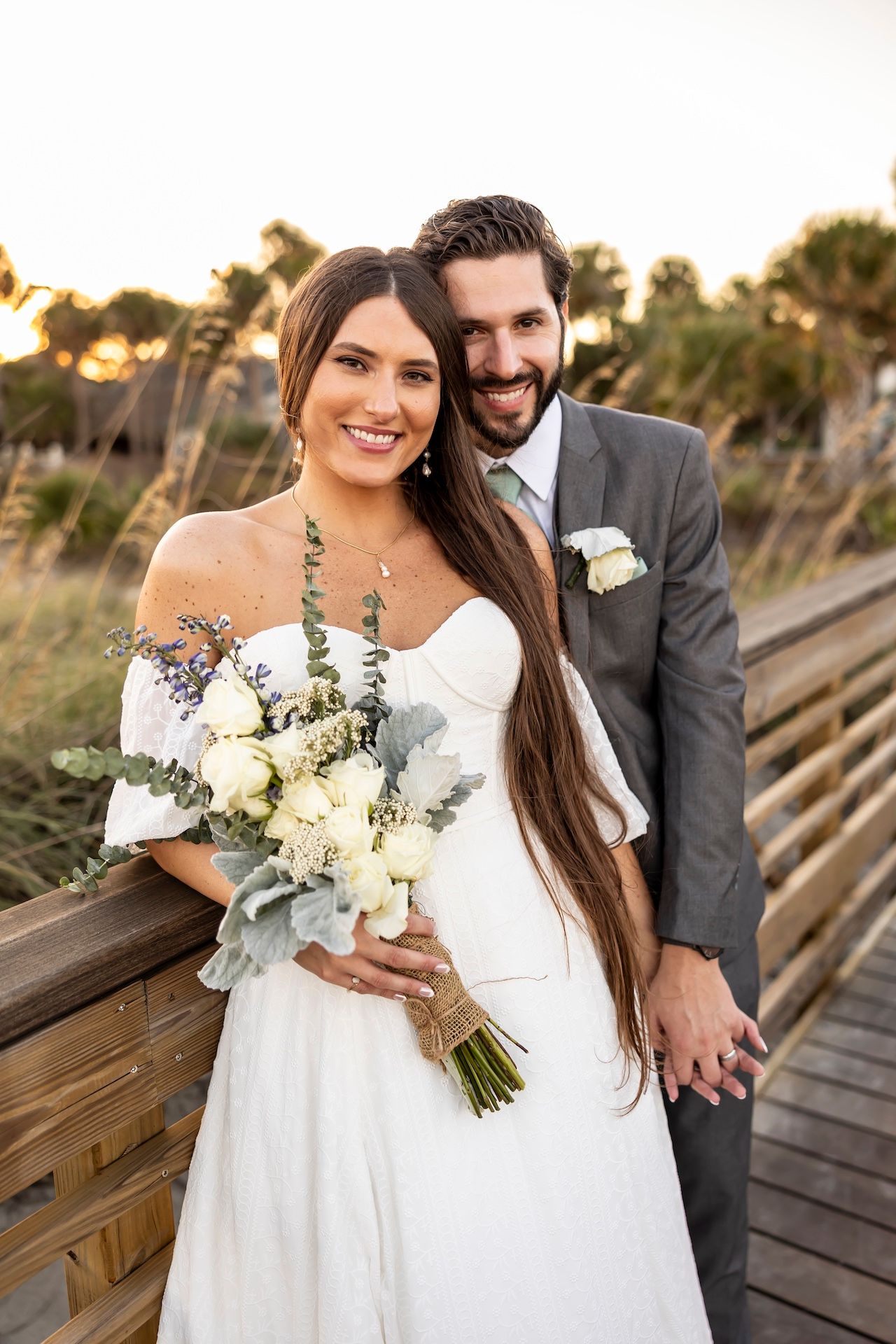 A happy couple poses on a wooden boardwalk at sunset, with the bride holding a white and green floral bouquet.