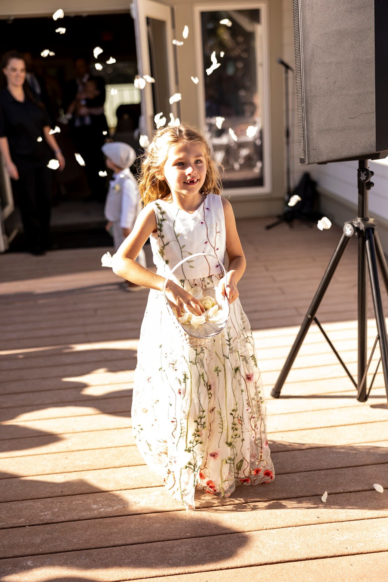 Young child in white floral dress dances joyfully in sunlit room with flower petals falling around her.