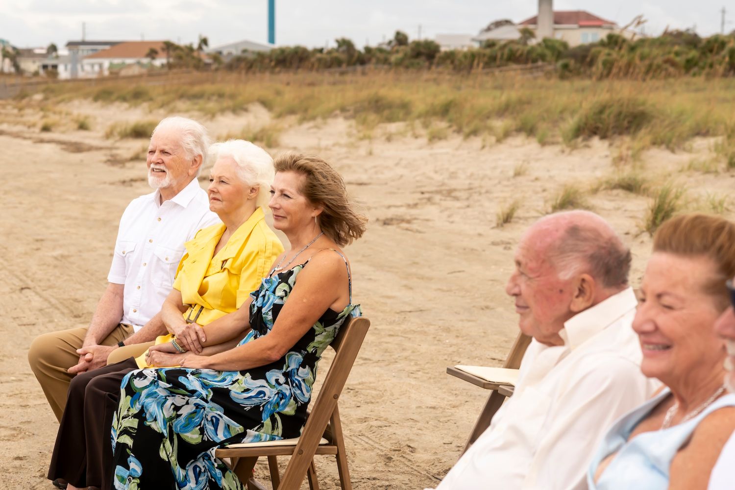 A group of senior friends laughing together while sitting in chairs on a sandy beach.