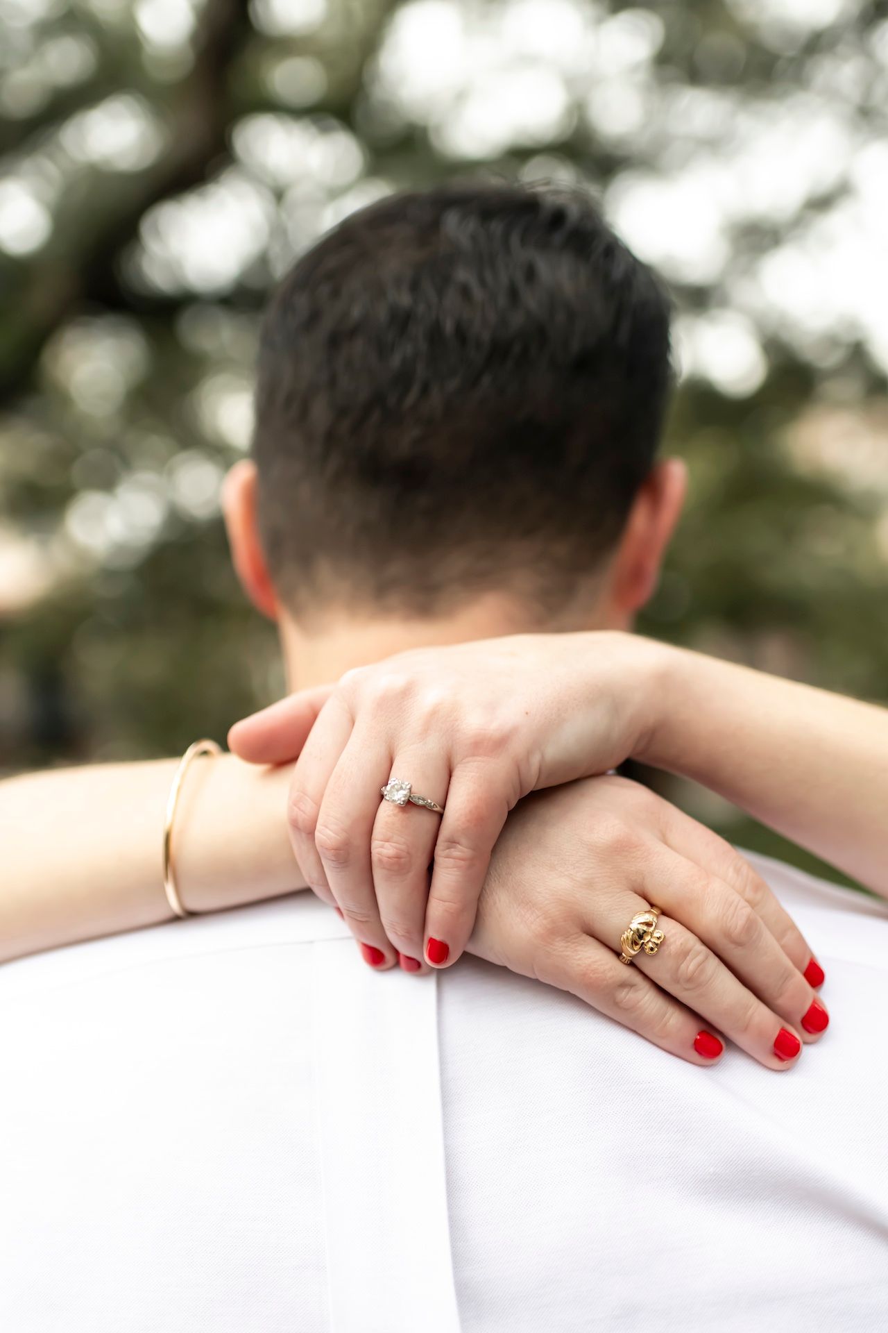Close-up of hands with wedding rings and red nail polish resting on a white surface with trees in background.