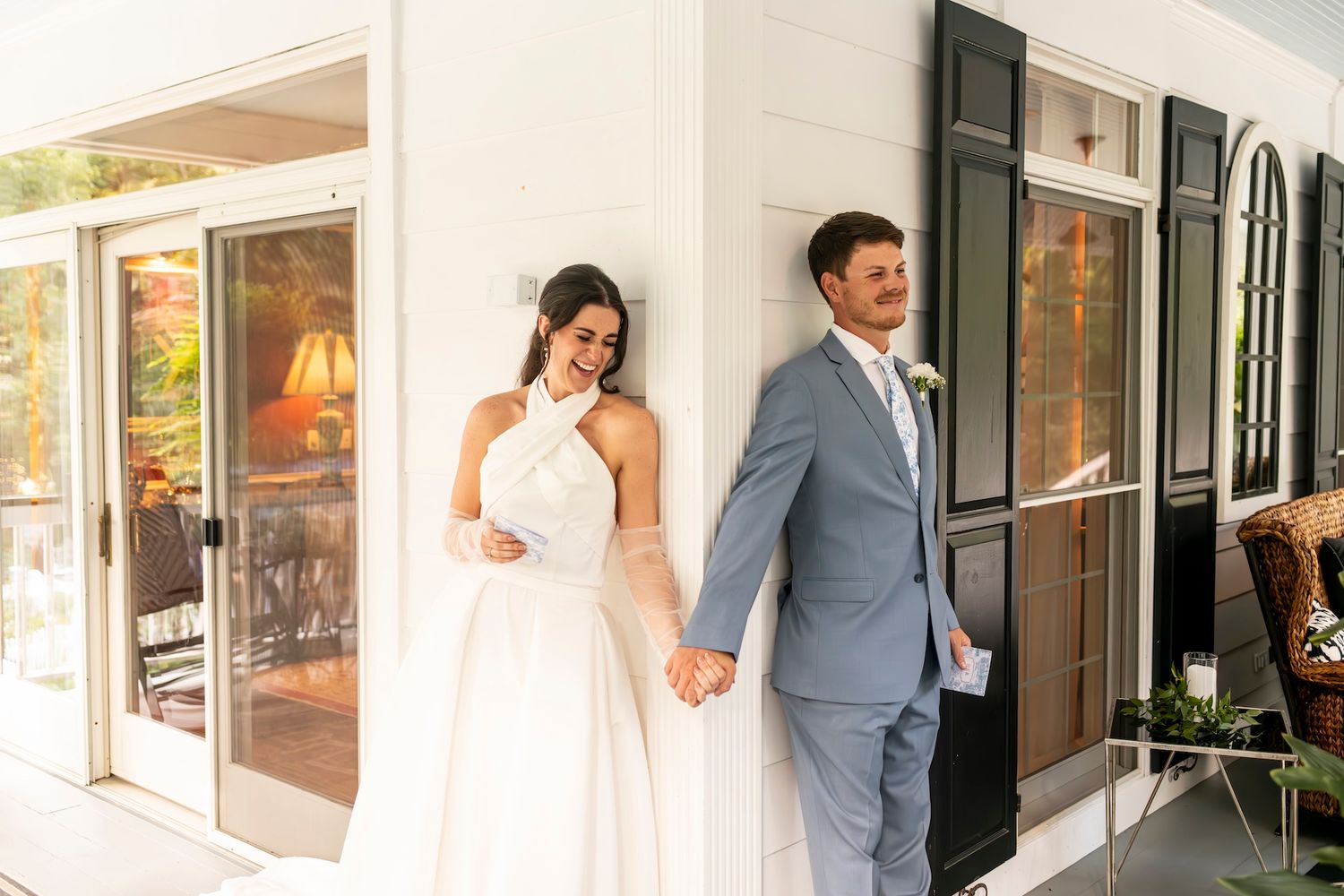 Newlywed couple holds hands in front of white building with black shutters on sunny autumn day.