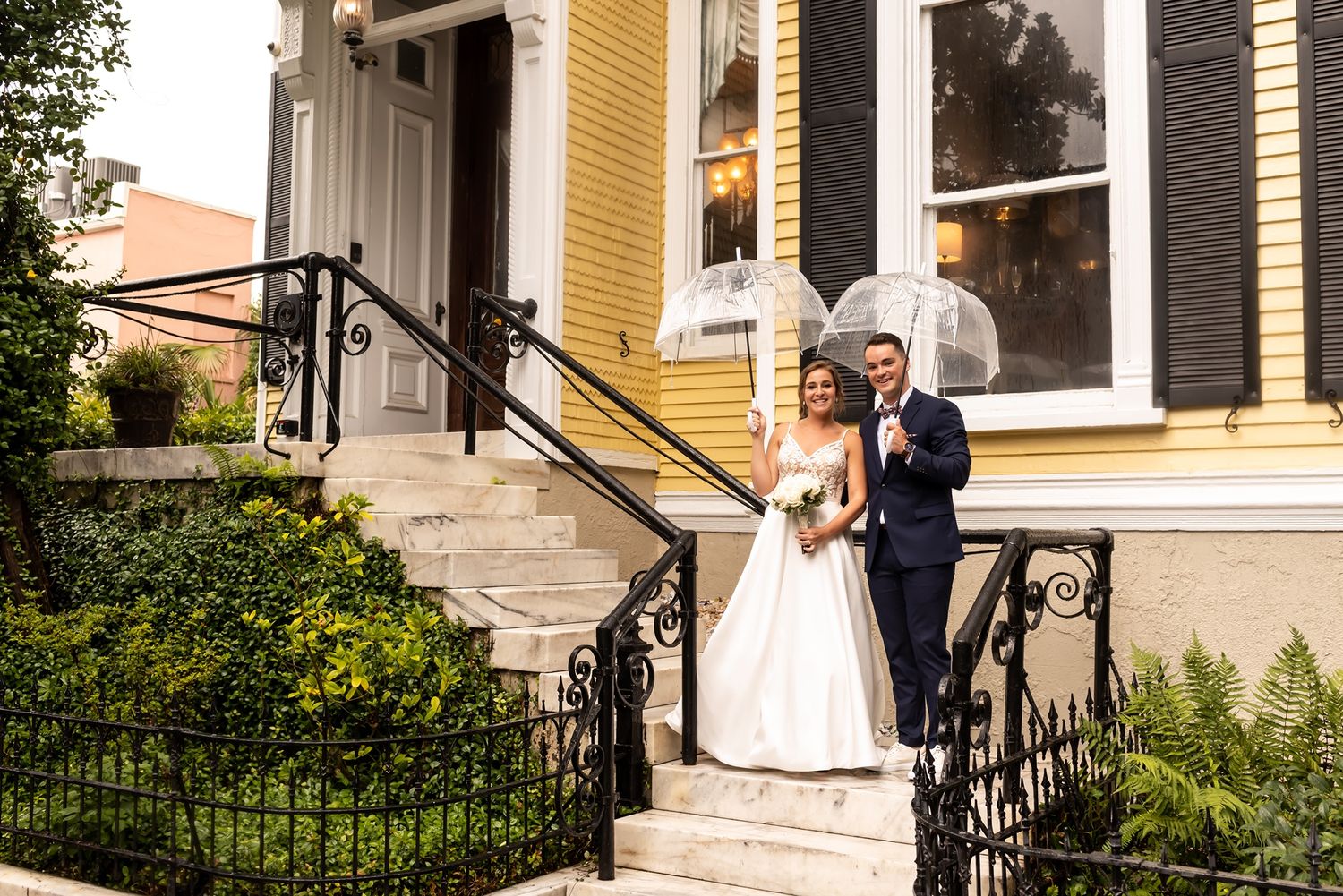 A couple in wedding attire stands on the steps of a yellow historic house with ornate iron railings.