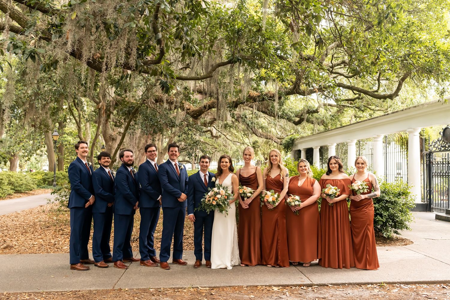 A wedding party stands under moss-draped oak trees, with bridesmaids in burgundy dresses and groomsmen in navy suits.