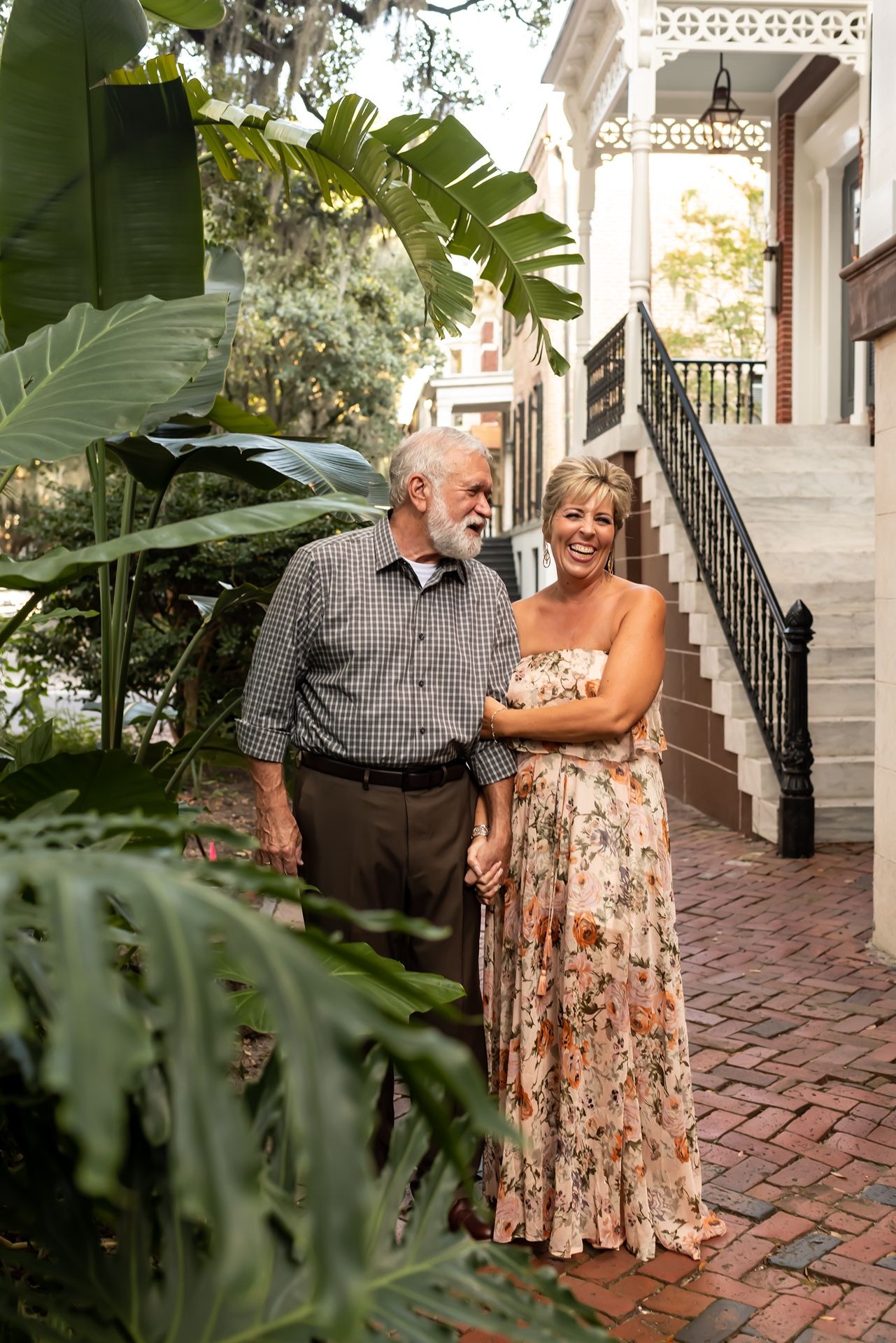 An elderly couple share playful and affectionate moments on a brick patio surrounded by tropical foliage.