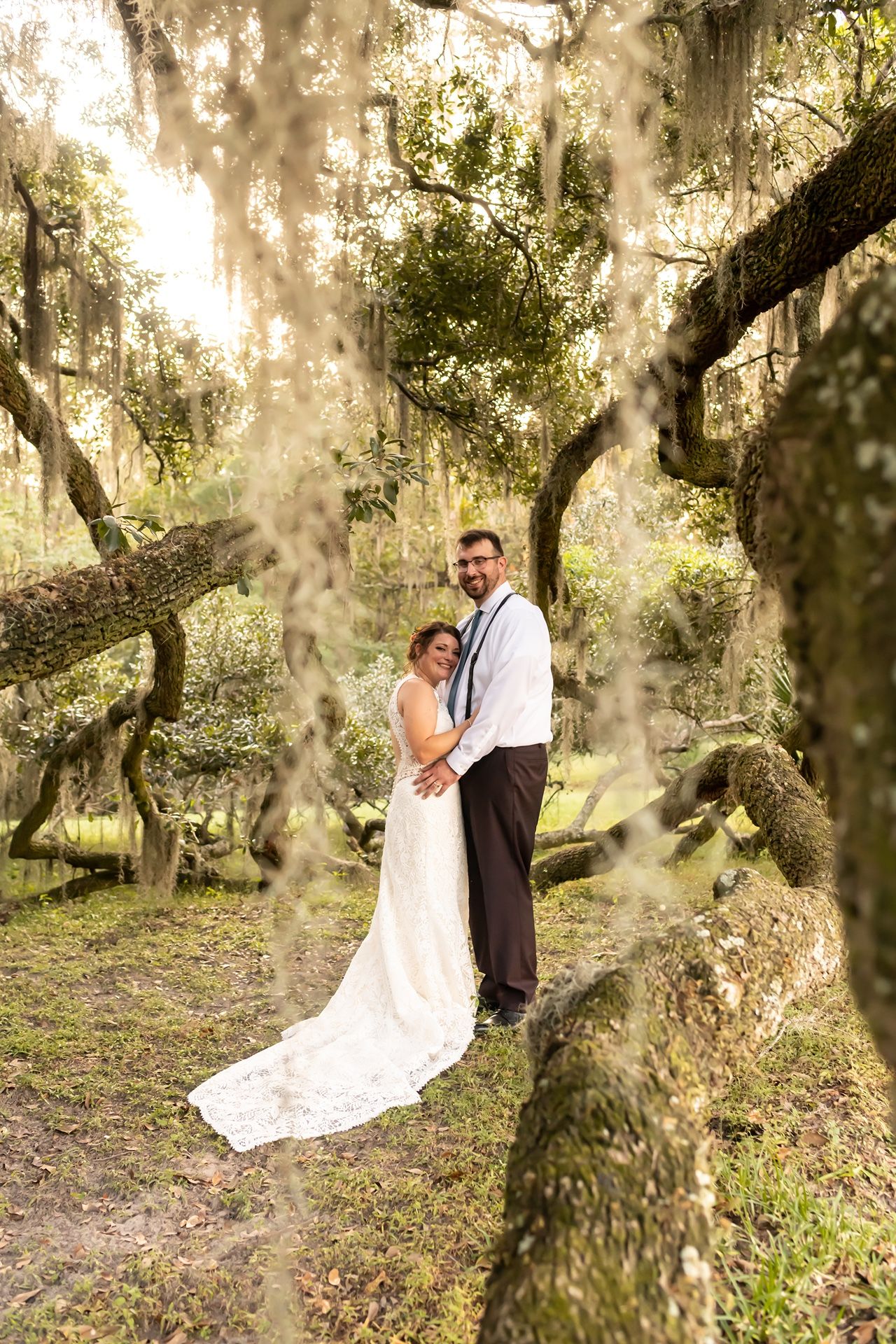 A couple in formal wedding attire embraces beneath moss-draped oak trees in a dreamy forest setting.