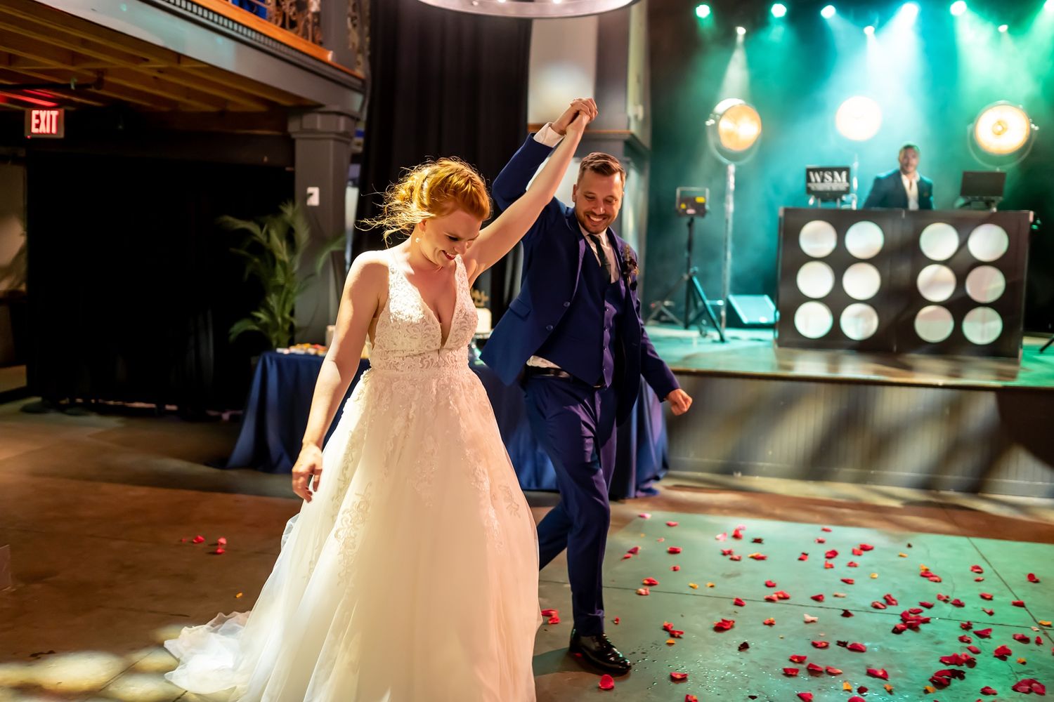 Two people dance together during a wedding celebration on a rose petal-covered floor with green stage lighting in the background.