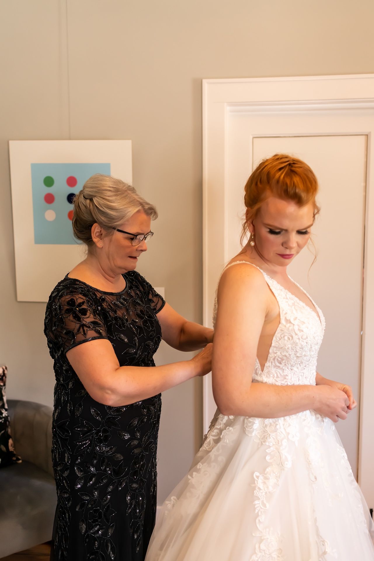 A person in a black lace dress helps adjust a white wedding gown in an indoor setting with framed artwork on the wall.