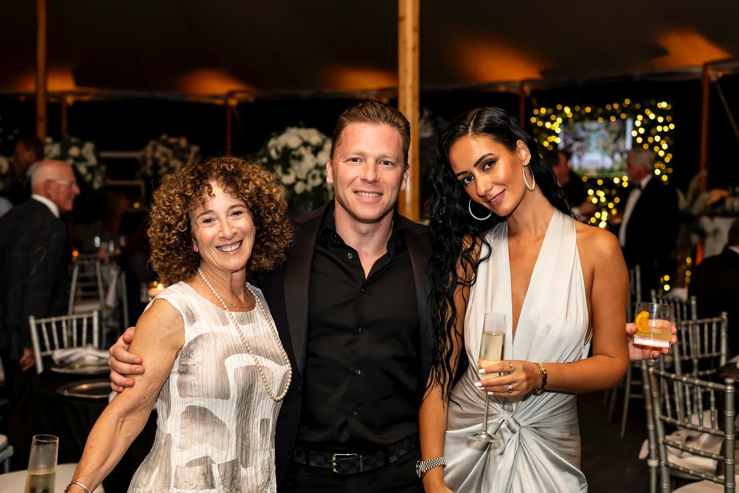 Three people in elegant evening attire pose together at a festive nighttime event with decorative lights in background.