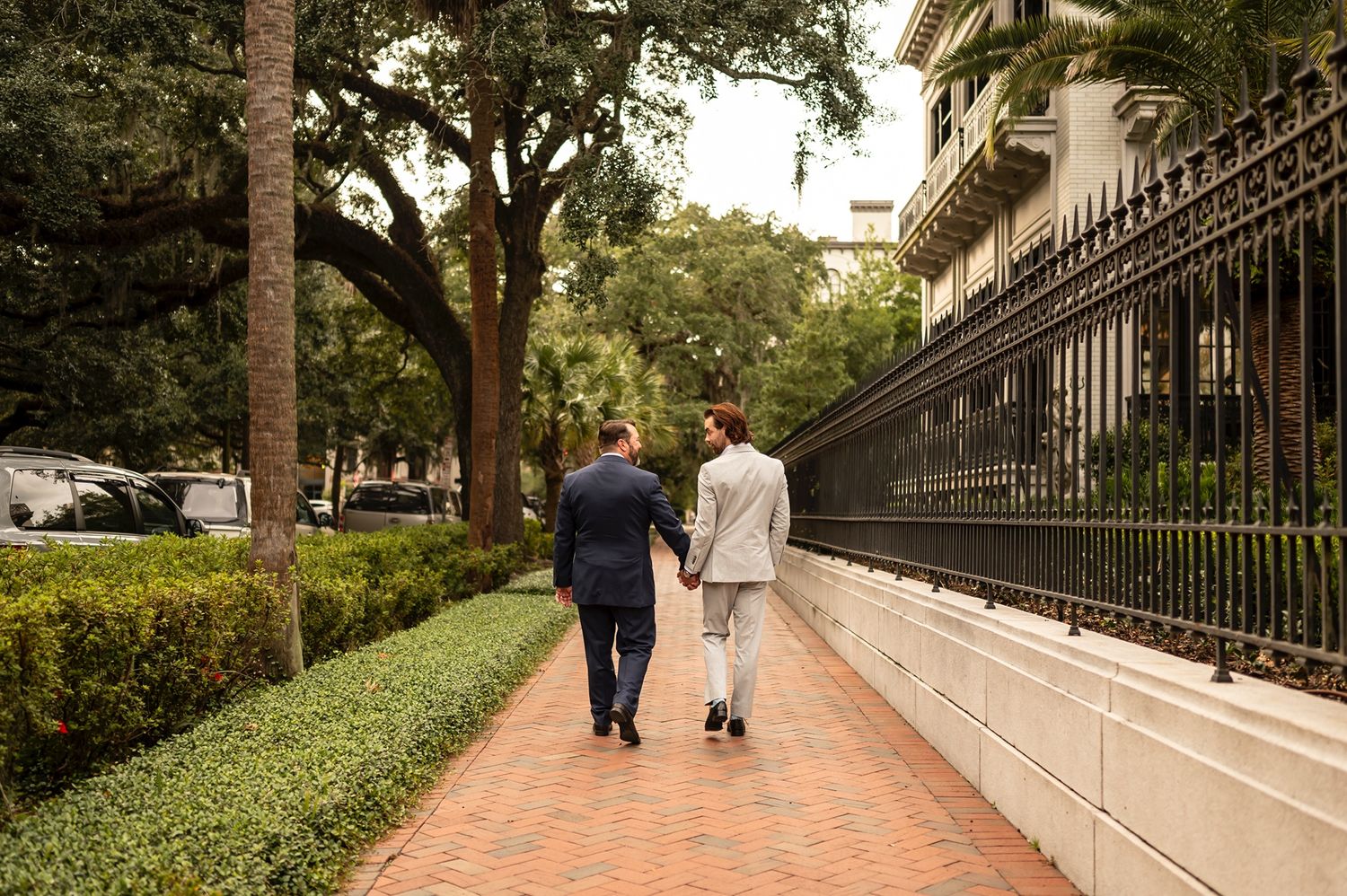 Two people walk along a brick pathway beside an ornate iron fence and lush greenery on a sunny afternoon.