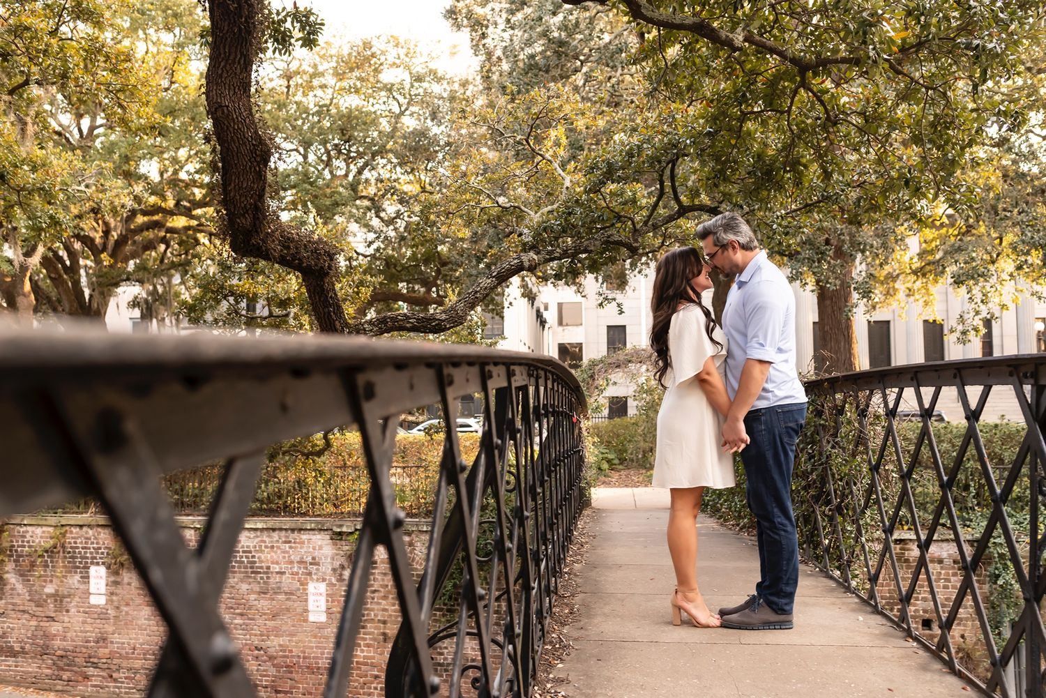 A couple shares a romantic moment on a rustic bridge path surrounded by autumn trees in a park setting.