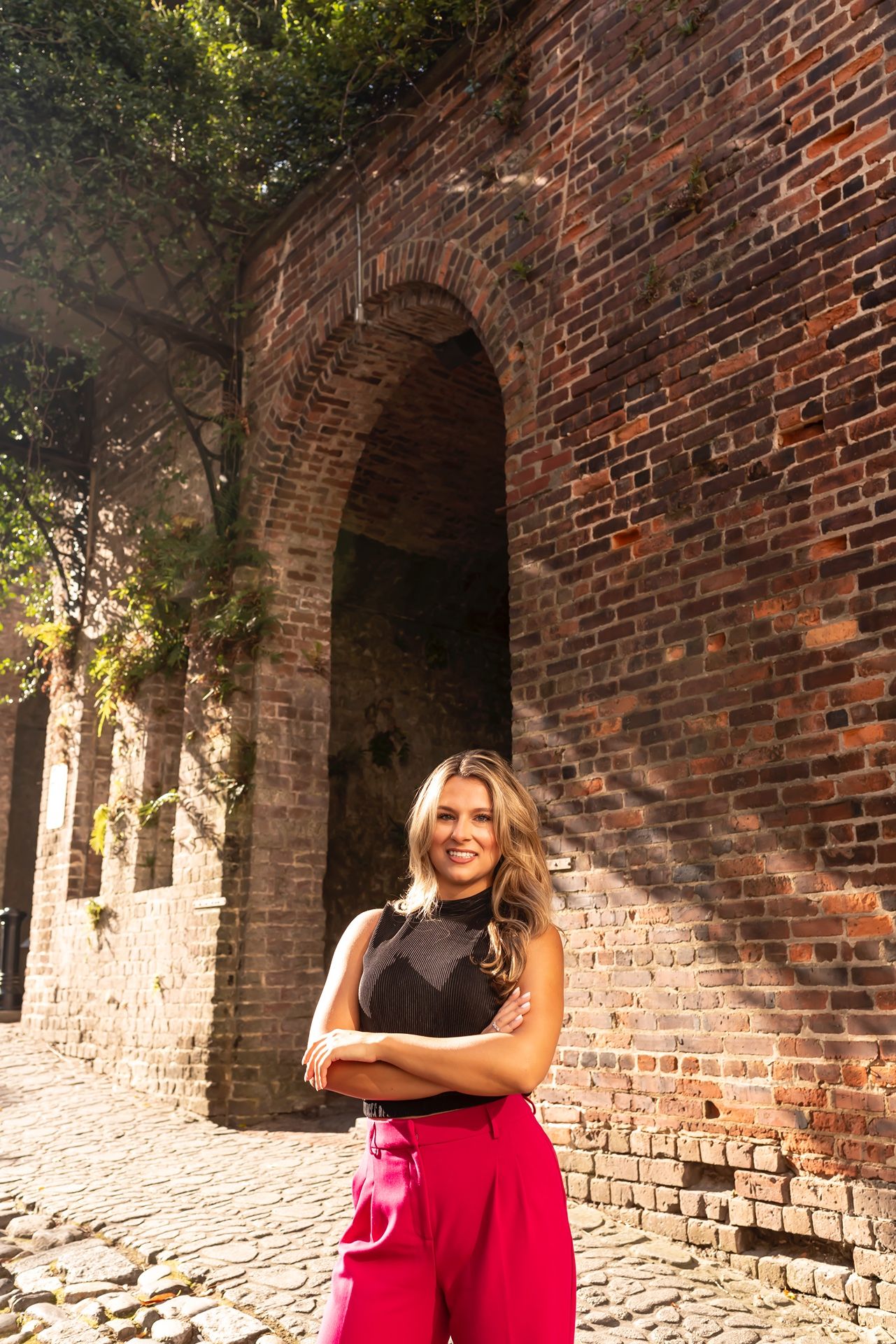 Person in casual attire standing in front of historic brick archway with sunlit cobblestone path.