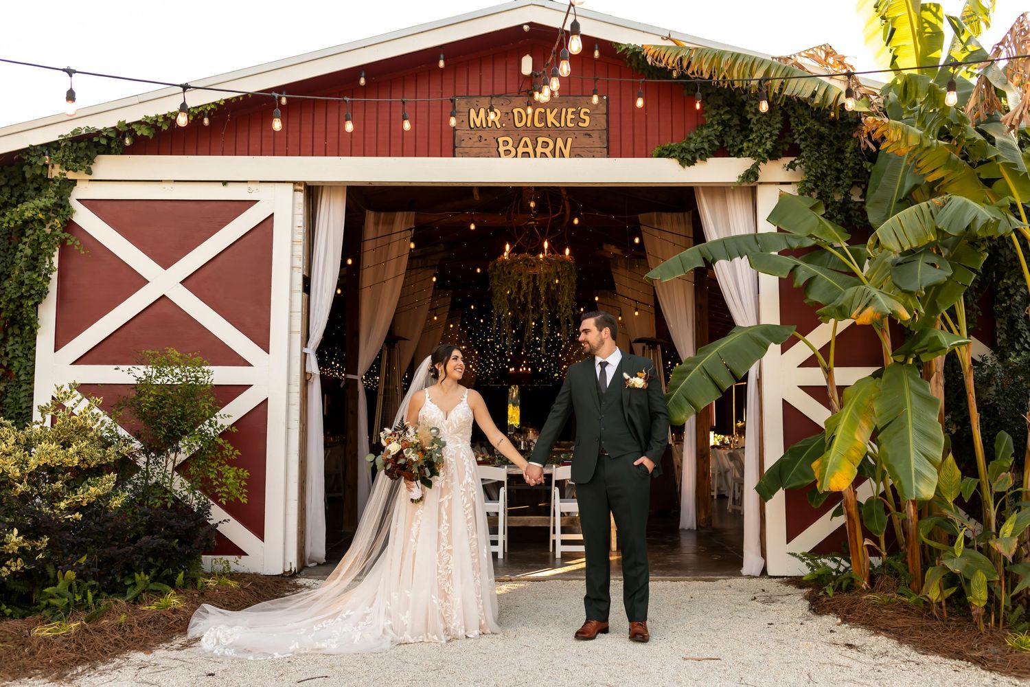 A bride and groom stands in front of a rustic red barn with white trim and tropical plants.