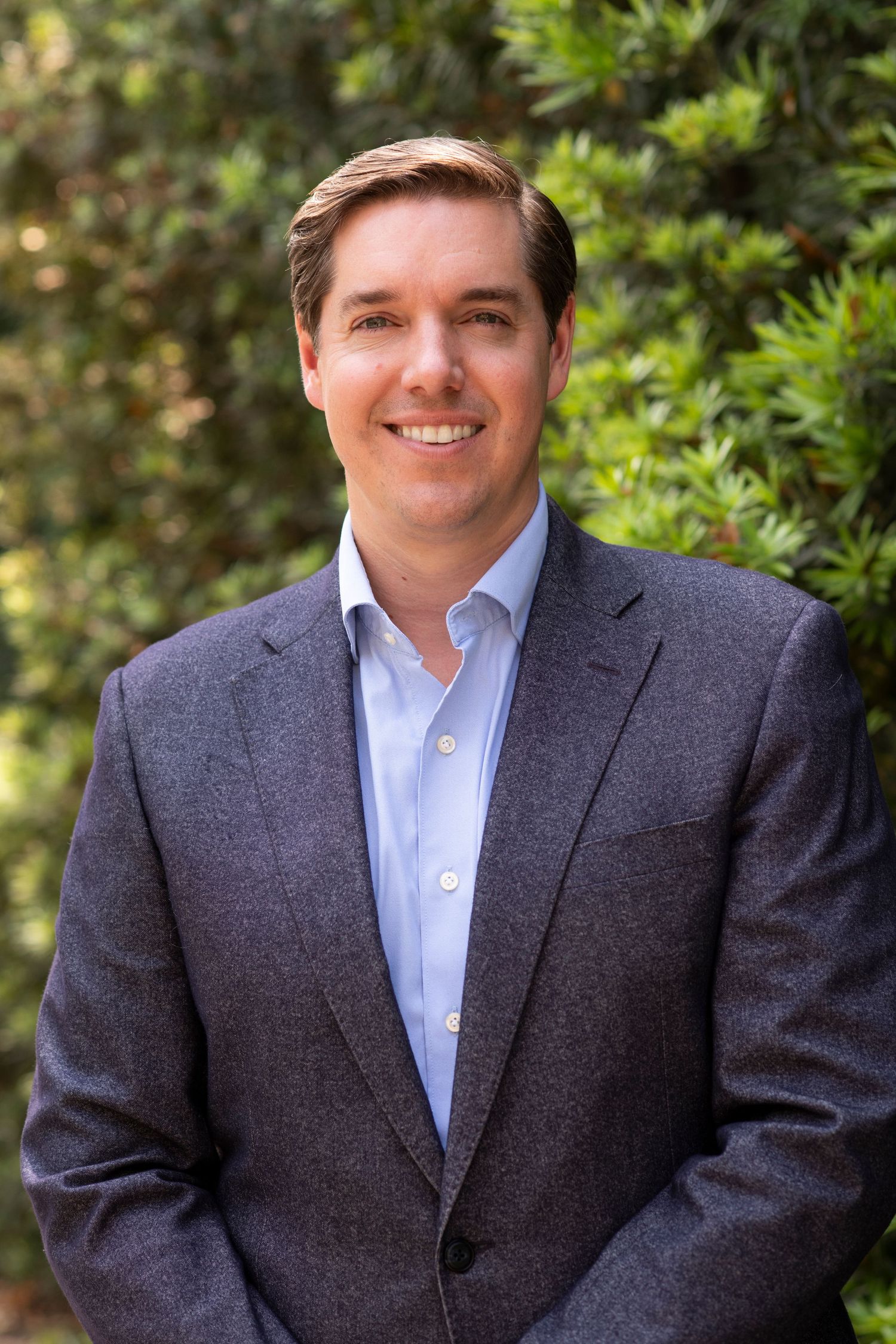 Professional headshot against green foliage background showing confident person in grey suit and light blue shirt.