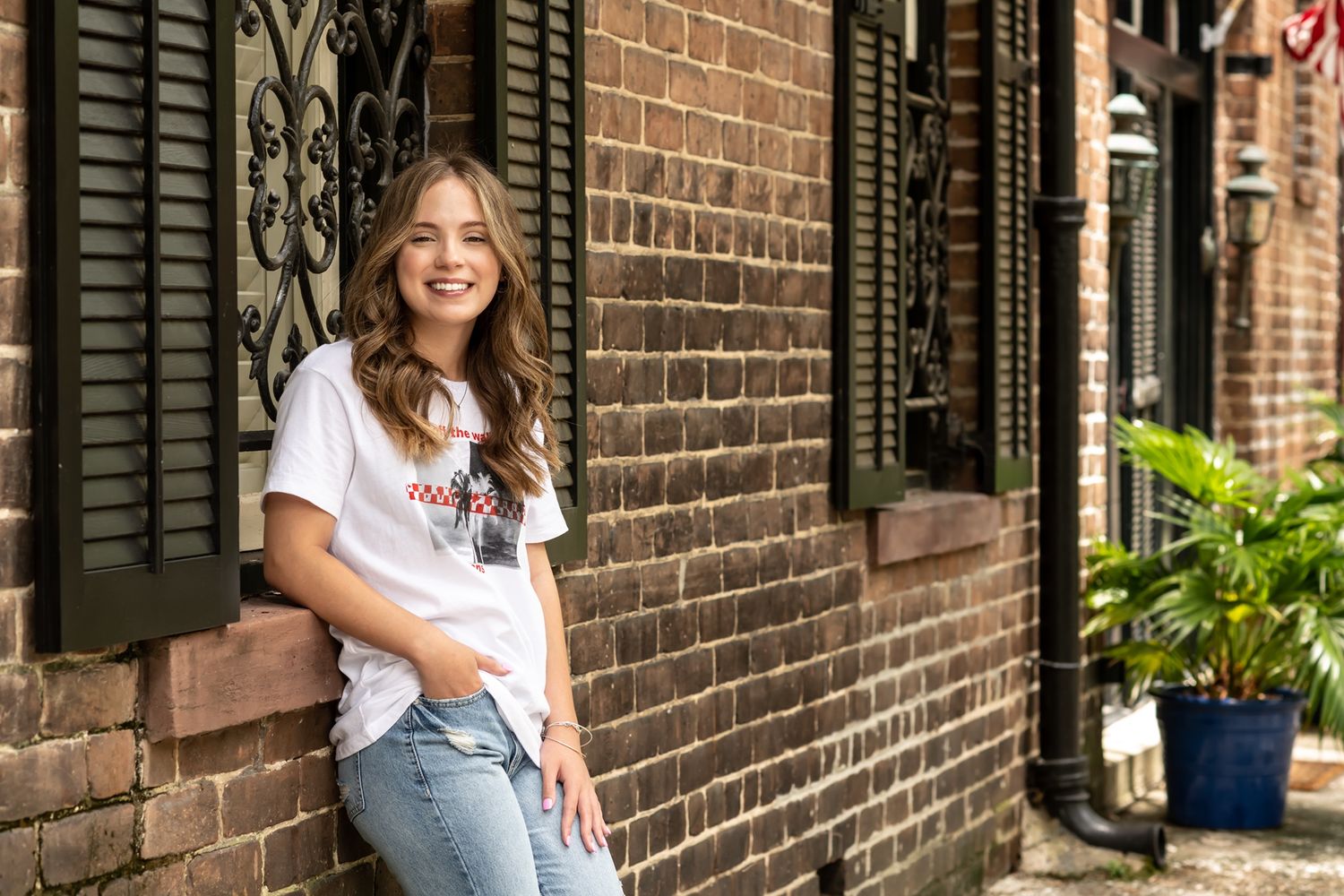 A smiling person in casual attire stands against a brick wall with black shutters on a sunny day in the city.
