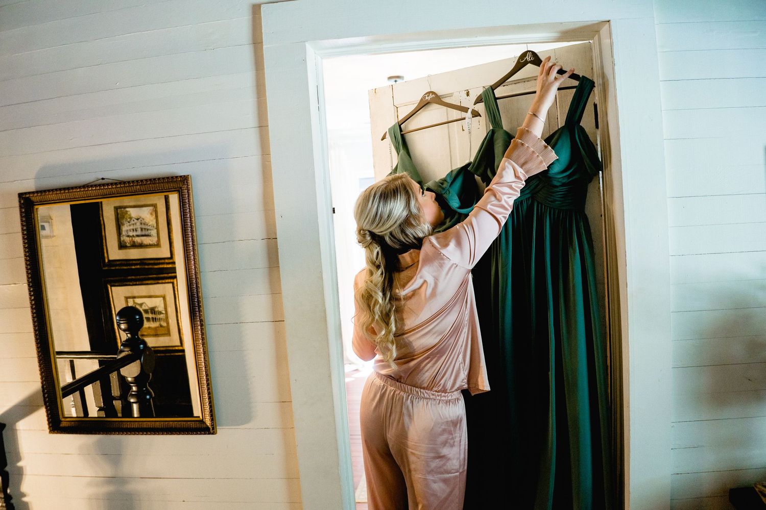 A person adjusts a dark green bridesmaid dress hanging on a closet door while a mirror reflects the scene.