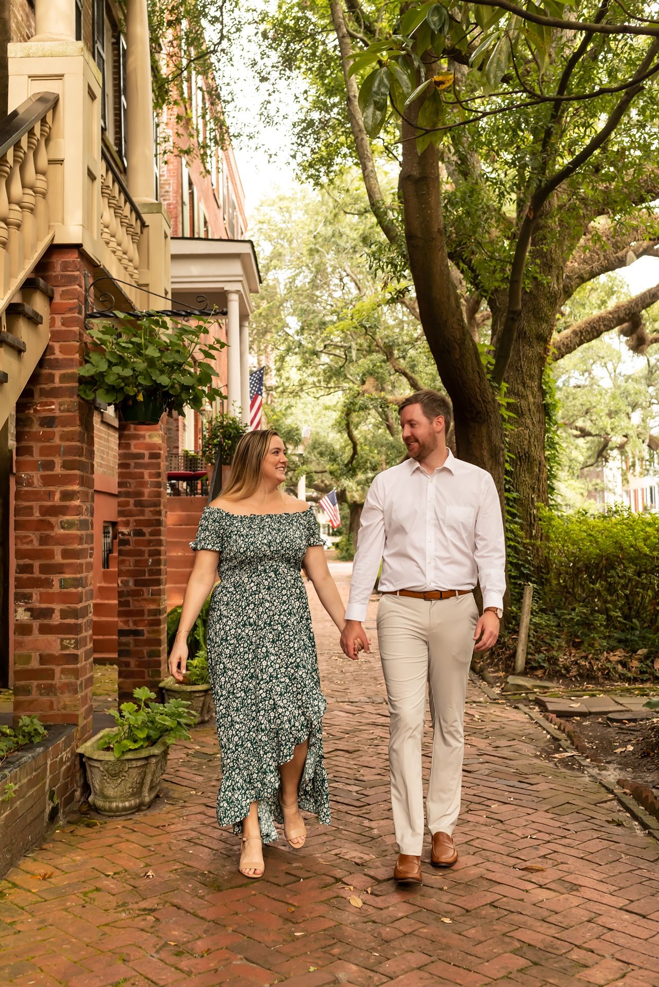 A couple walks hand in hand down a charming brick sidewalk lined with historic buildings and lush greenery.