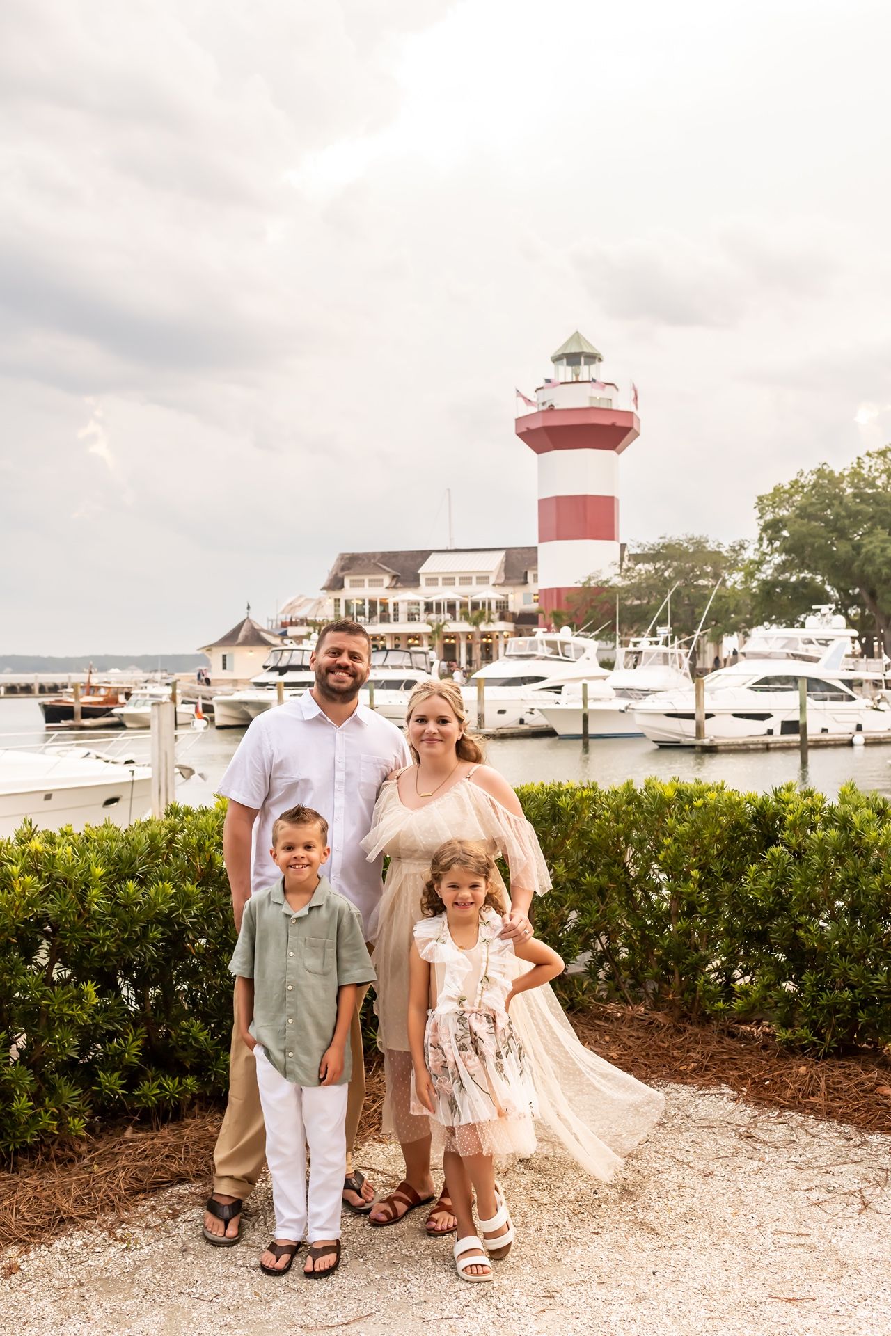 A family stands on a gravel path at a coastal marina with a red and white lighthouse tower in the background.