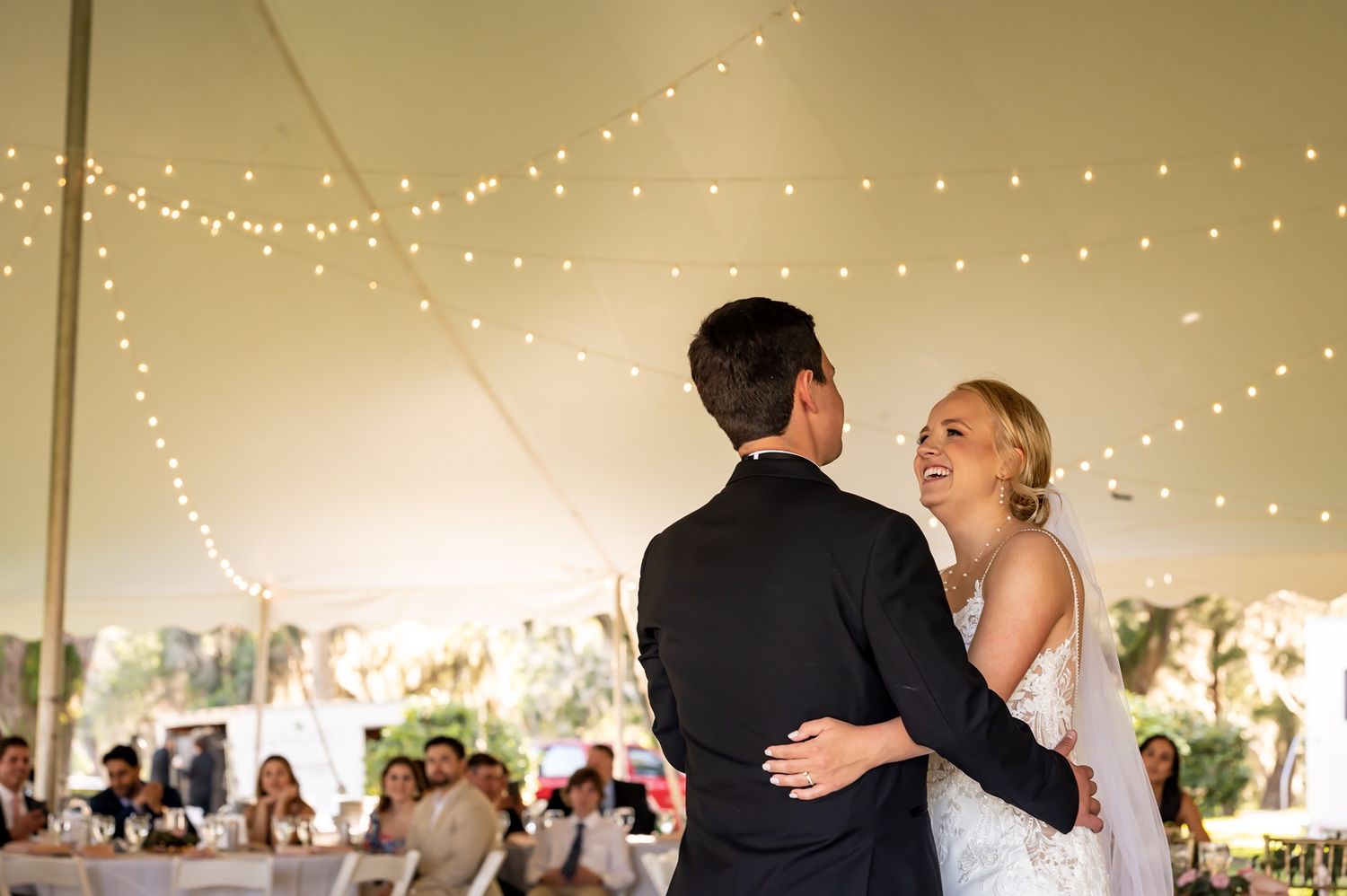 A couple share their first dance as newlyweds under romantic string lights in an elegant white wedding reception tent.