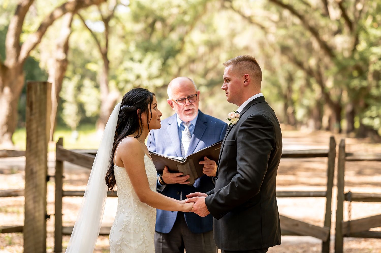 Wedding couple holds hands during outdoor ceremony under oak trees while officiant reads from book.