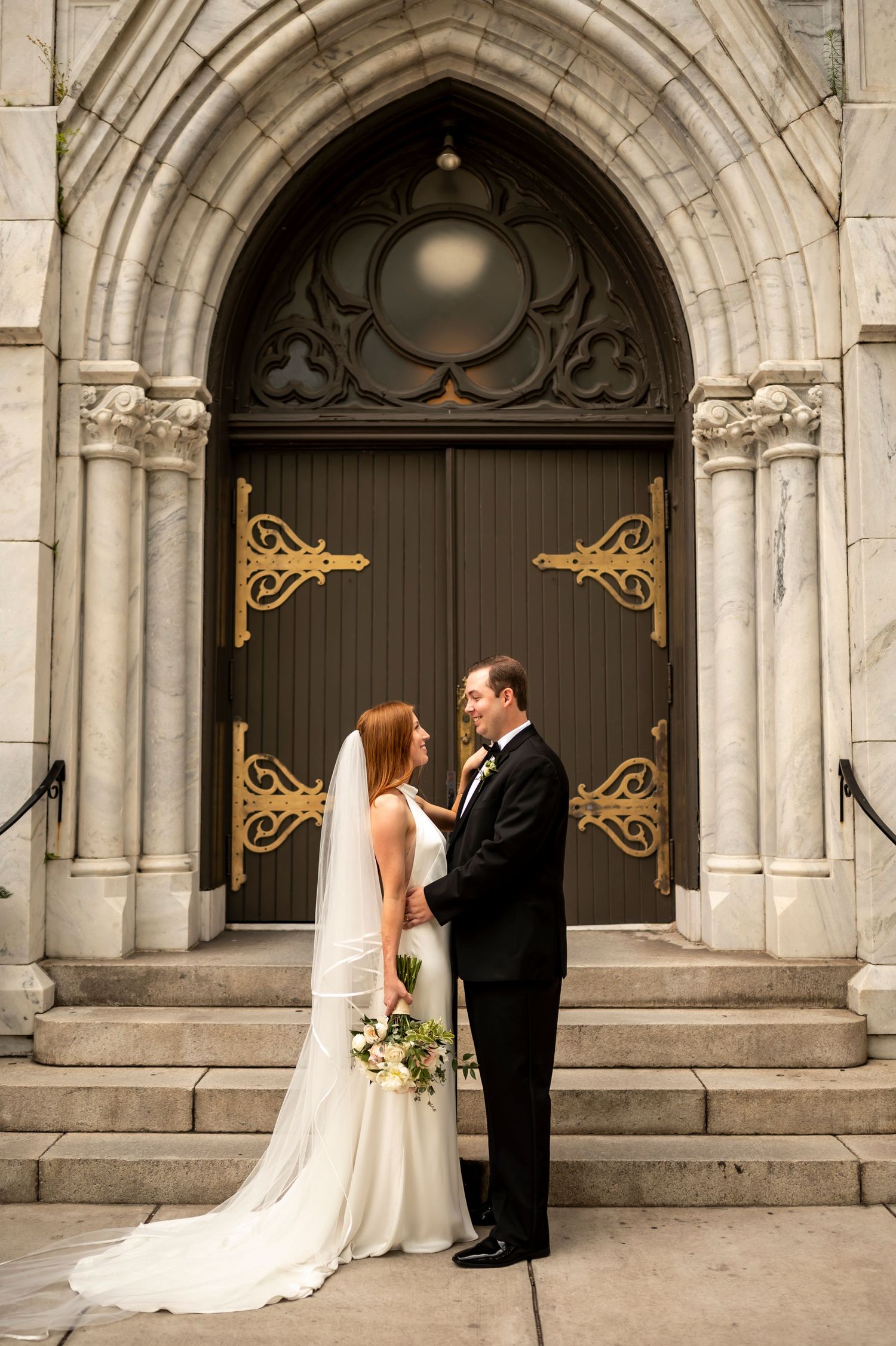 A wedding couple stands on stone steps in front of an ornate Gothic church door with decorative gold hinges.