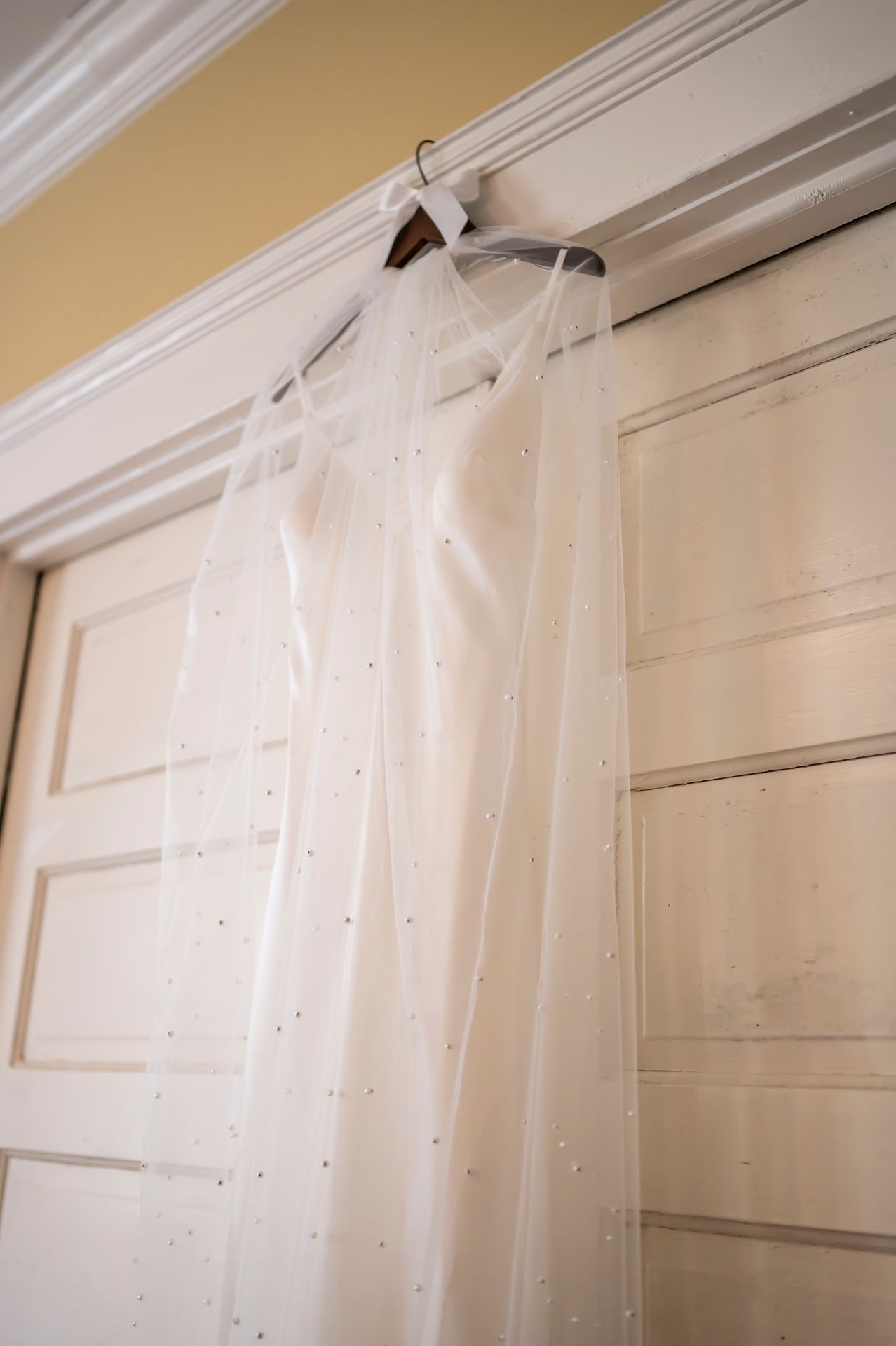 A delicate white wedding veil with sparkling rhinestone accents hangs elegantly against a cream-colored door.