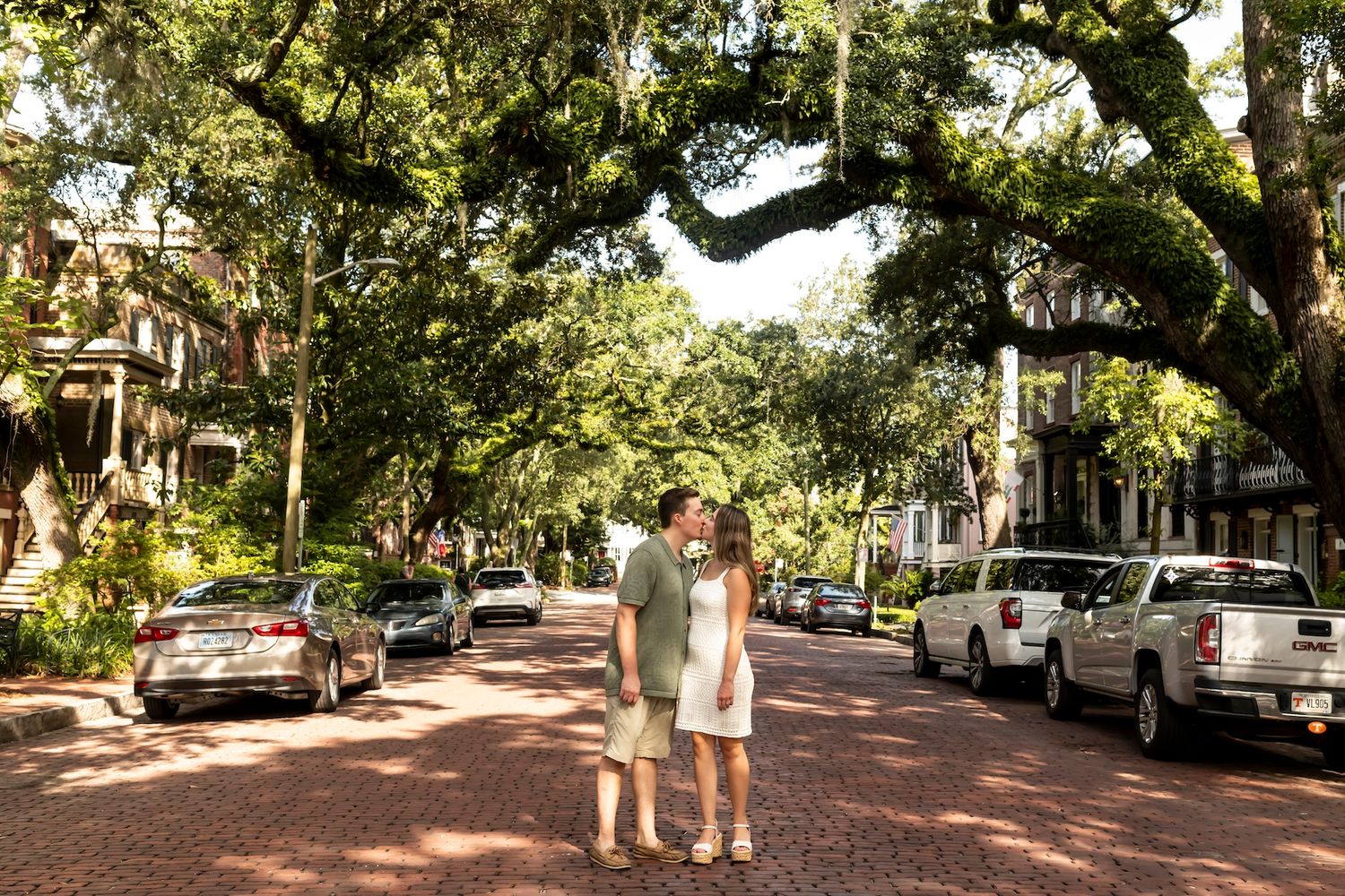 Two people stand on a brick-paved street lined with parked cars and oak trees draped in Spanish moss in a historic district.