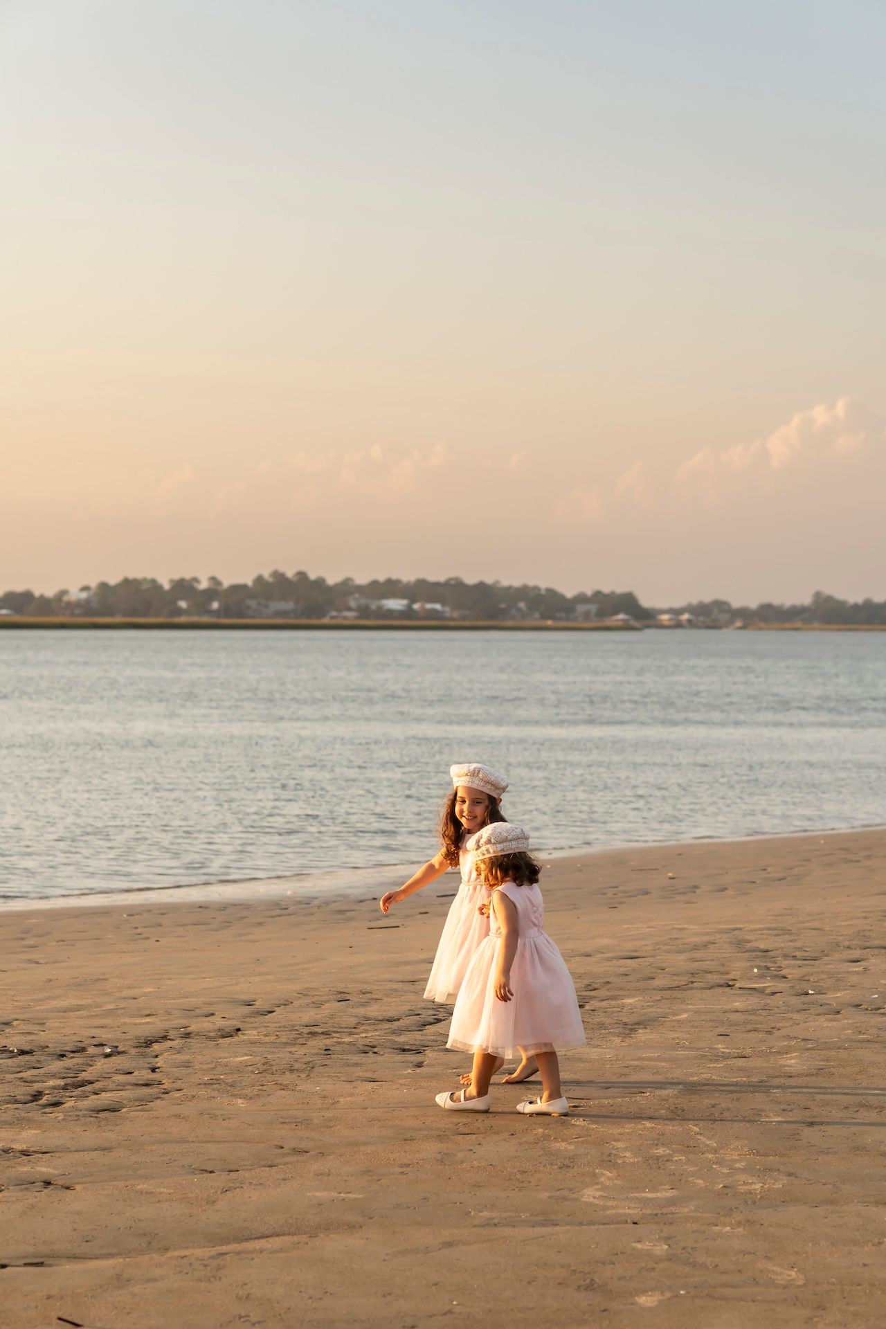 Two figures walk along a sandy beach at sunset with calm ocean waters and distant shoreline in golden light.