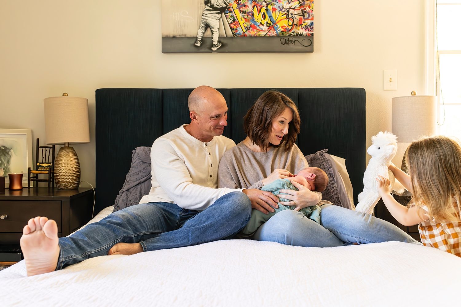 Family relaxes together on a bed with table lamps and colorful wall art visible in background.