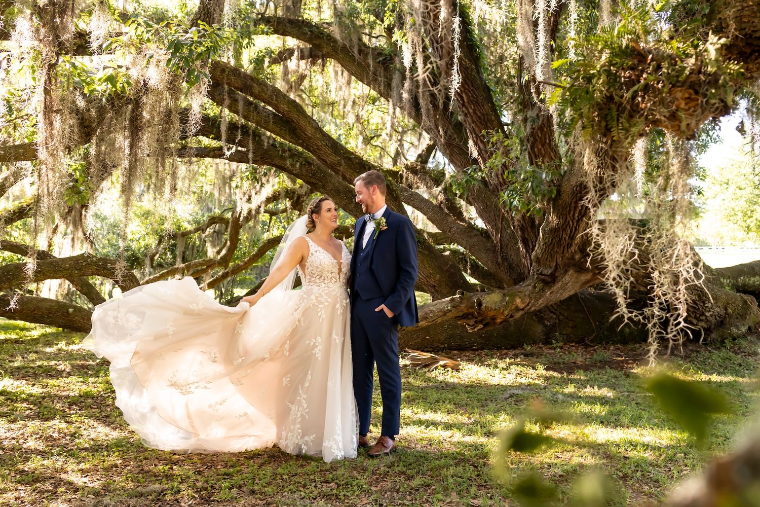 A romantic wedding photograph captured under a majestic oak tree draped with Spanish moss in a sunlit garden setting.