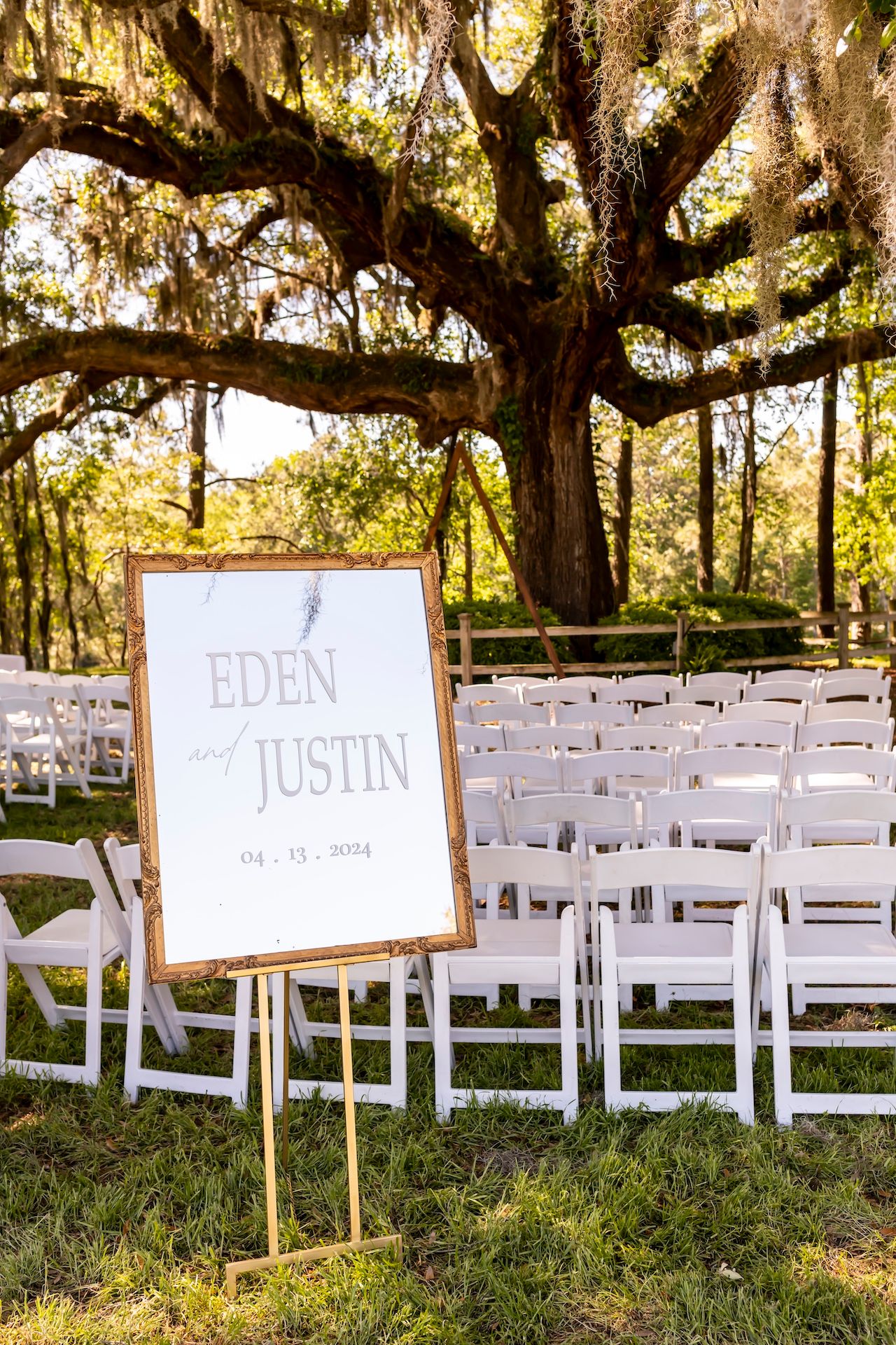 White folding chairs arranged under moss-draped oak trees for an outdoor wedding ceremony with rustic wooden name signs.