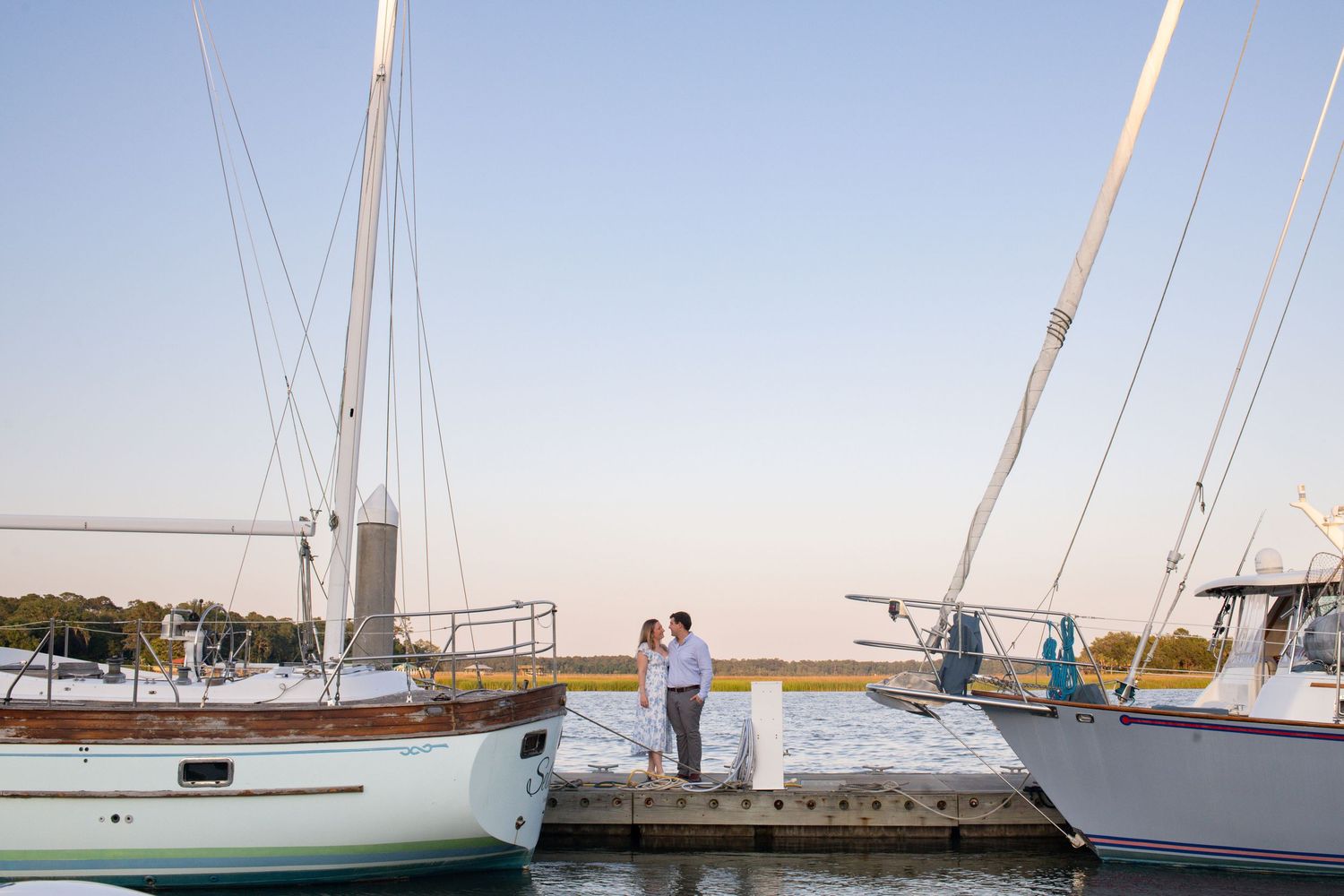A couple shares a romantic moment on a marina dock between two sailboats at sunset.