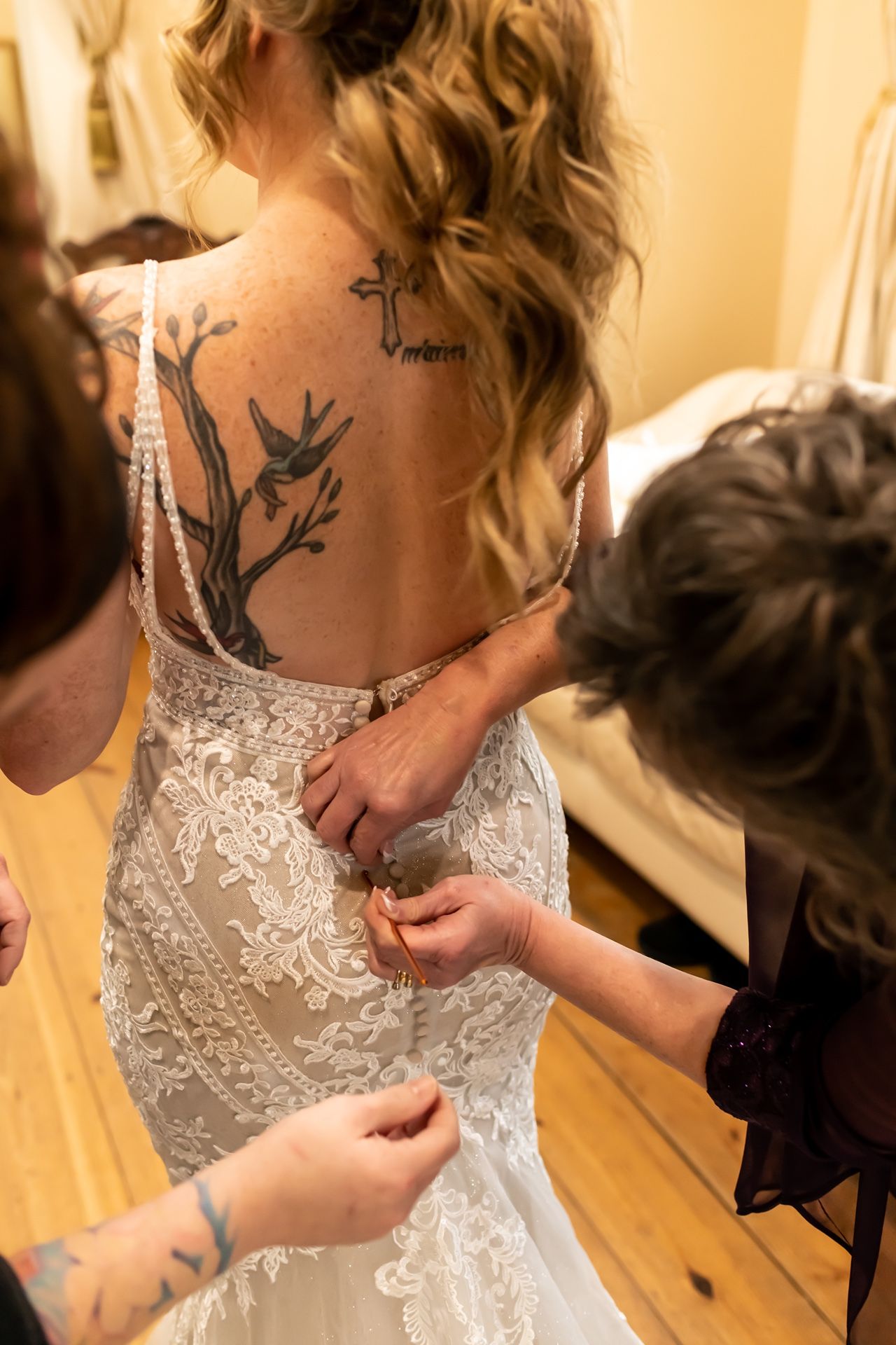 A close-up of someone being helped into a white lace wedding dress, showing an elegant back tattoo design.