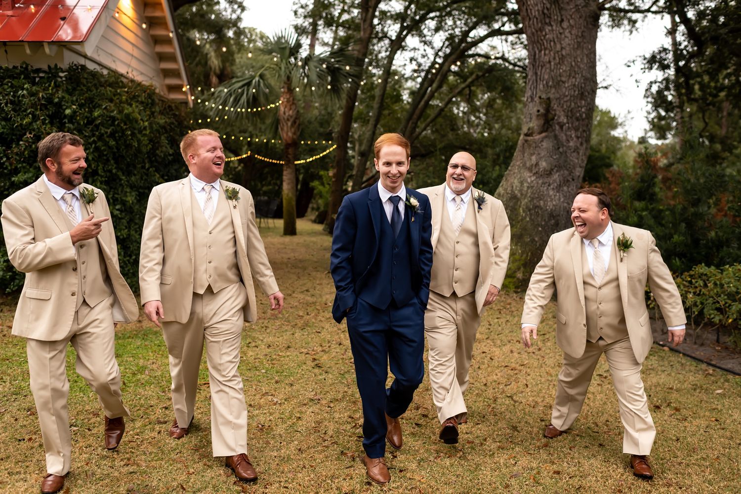 Groomsmen in beige suits walk and laugh with groom in navy blue suit on grassy lawn.