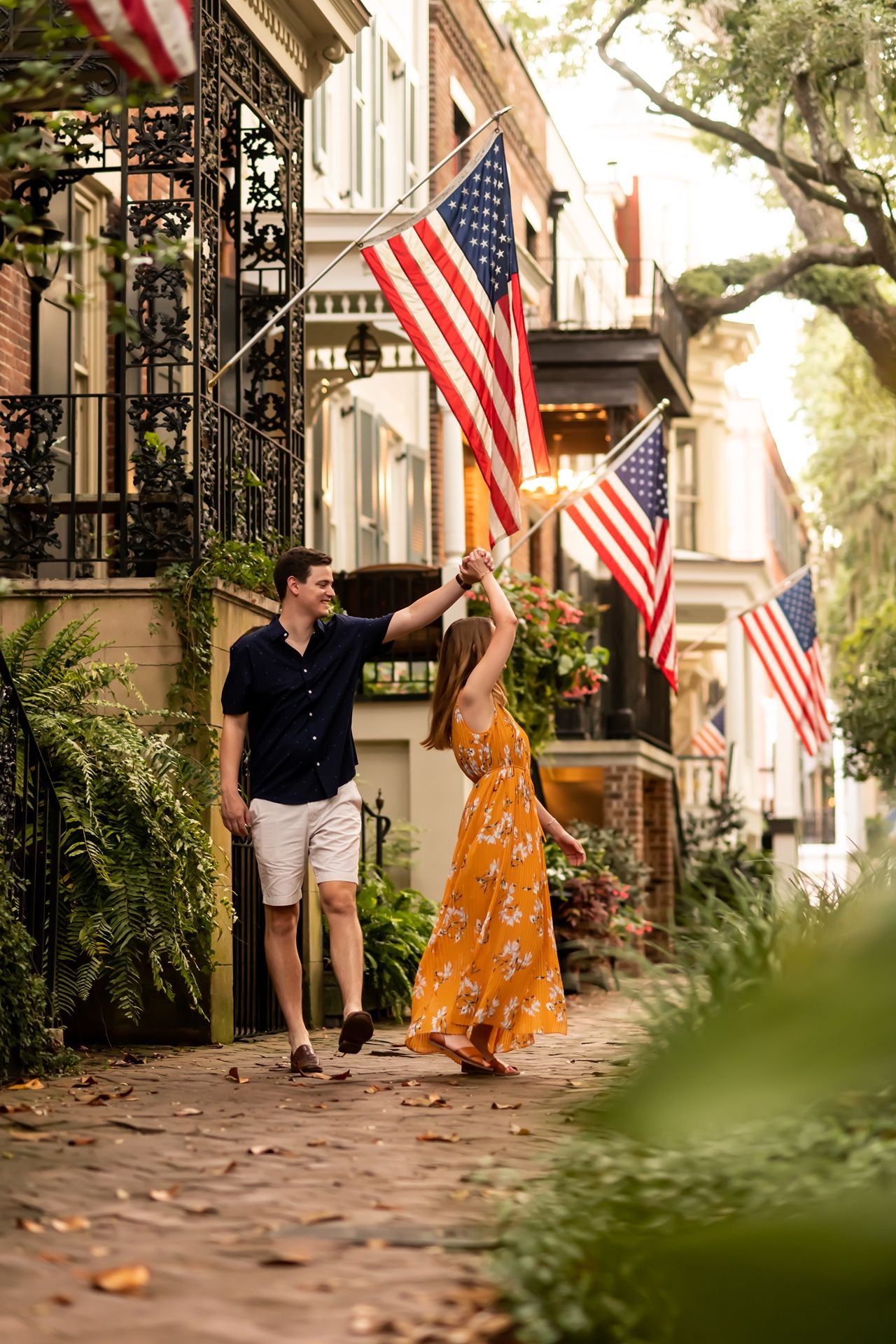 A romantic couple dances and spins on a tree-lined cobblestone street with American flags hanging from buildings.