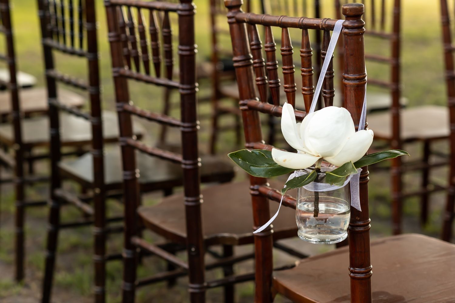 Dark wooden chairs in rows with white floral decorations set up for an outdoor wedding ceremony.