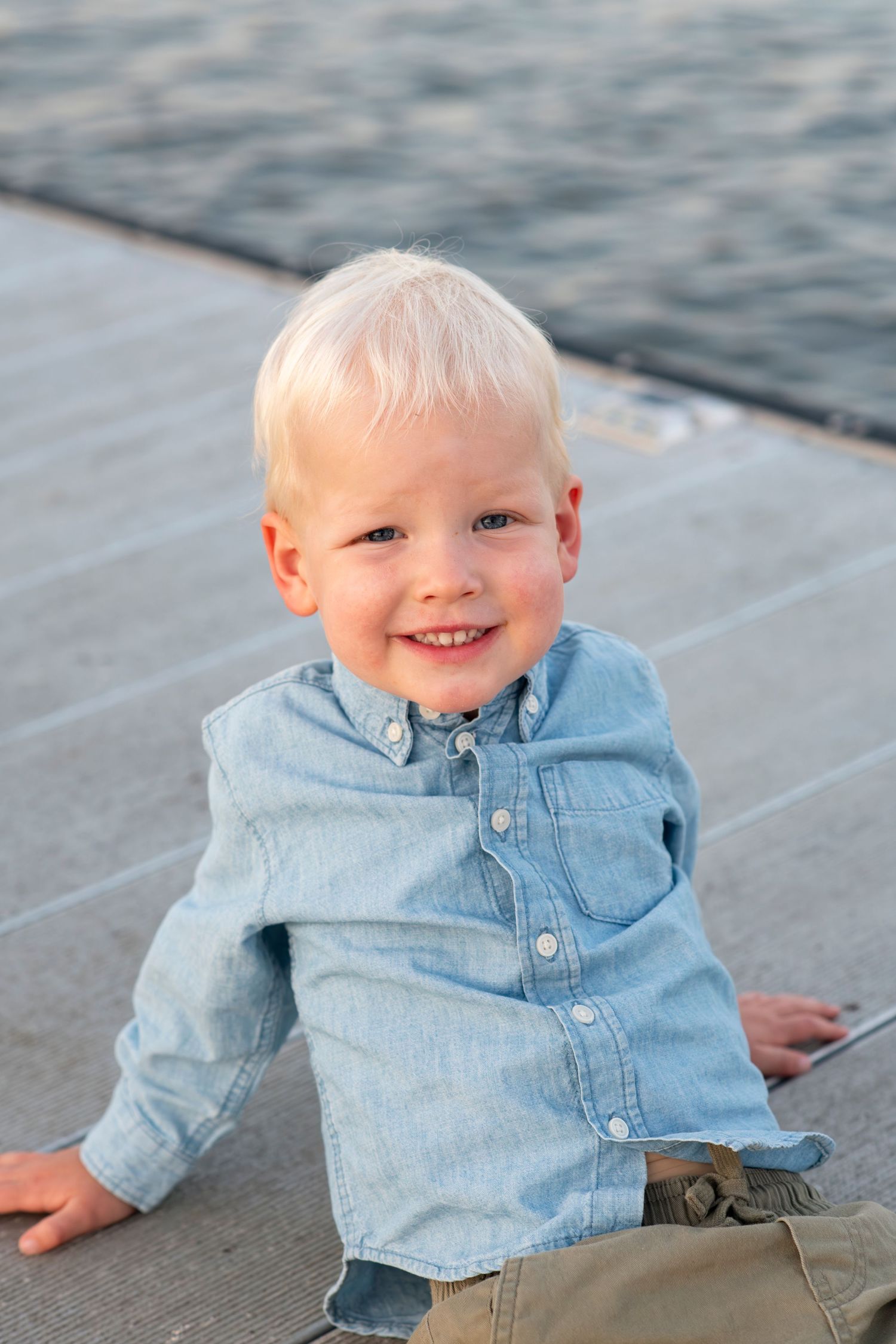 Smiling blonde toddler boy in a blue denim shirt sitting on a waterfront dock.
