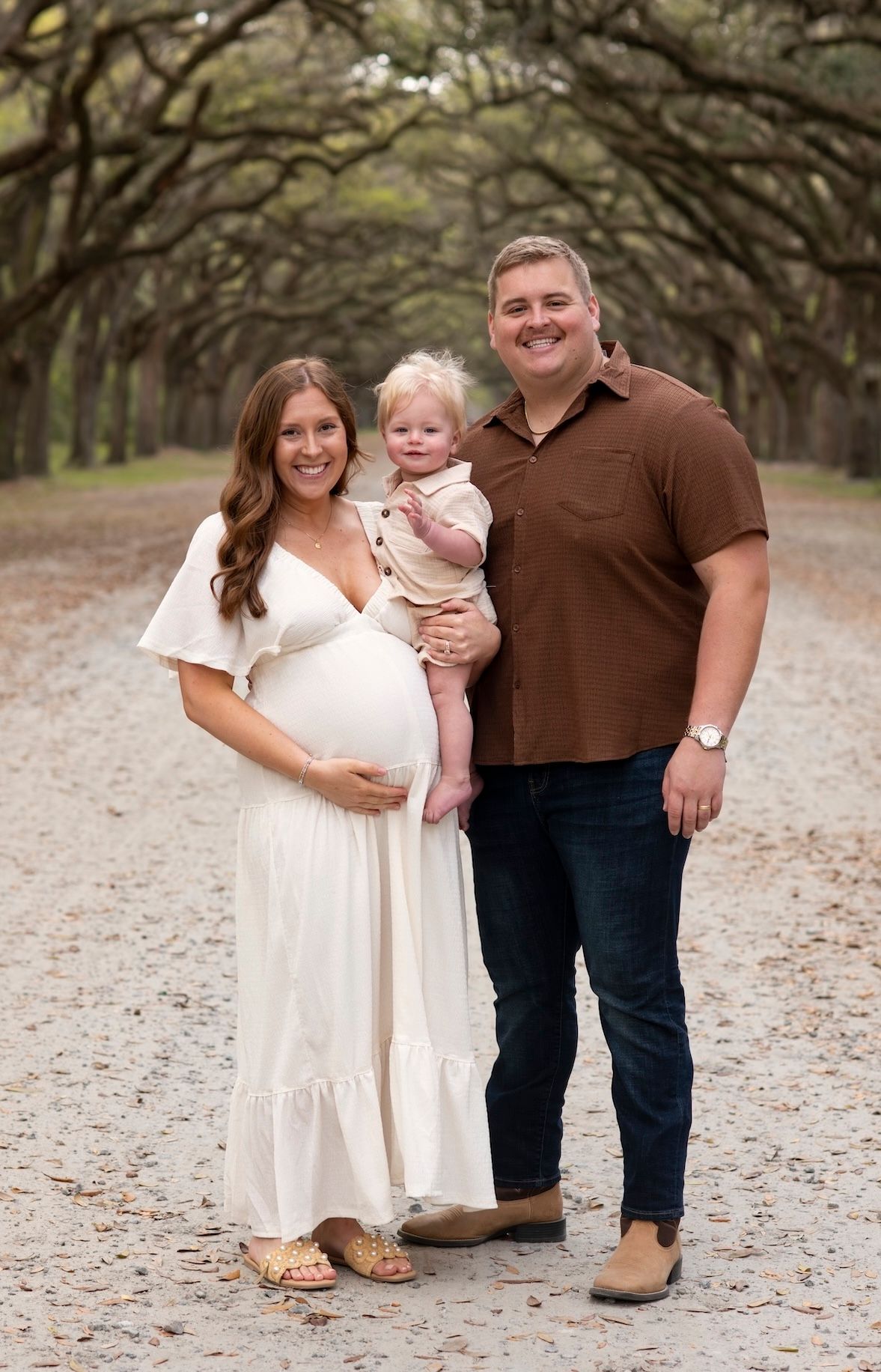 Pregnant woman in white dress poses with husband and toddler on a tree-lined gravel path for a maternity photo session.