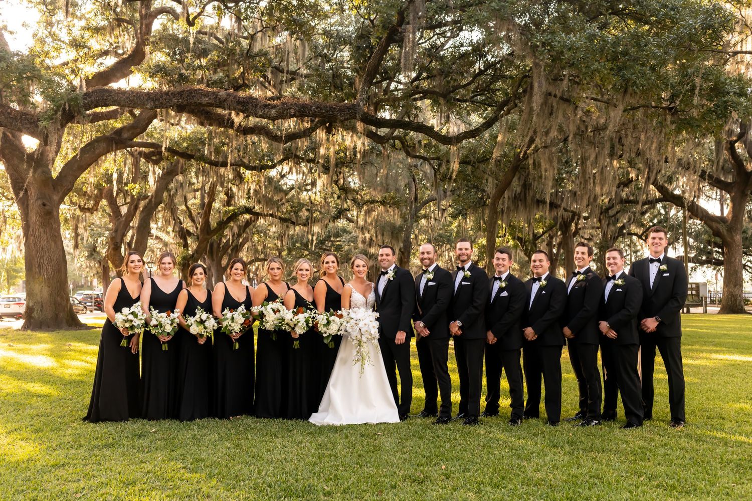 Wedding party in formal attire posing under large oak trees.