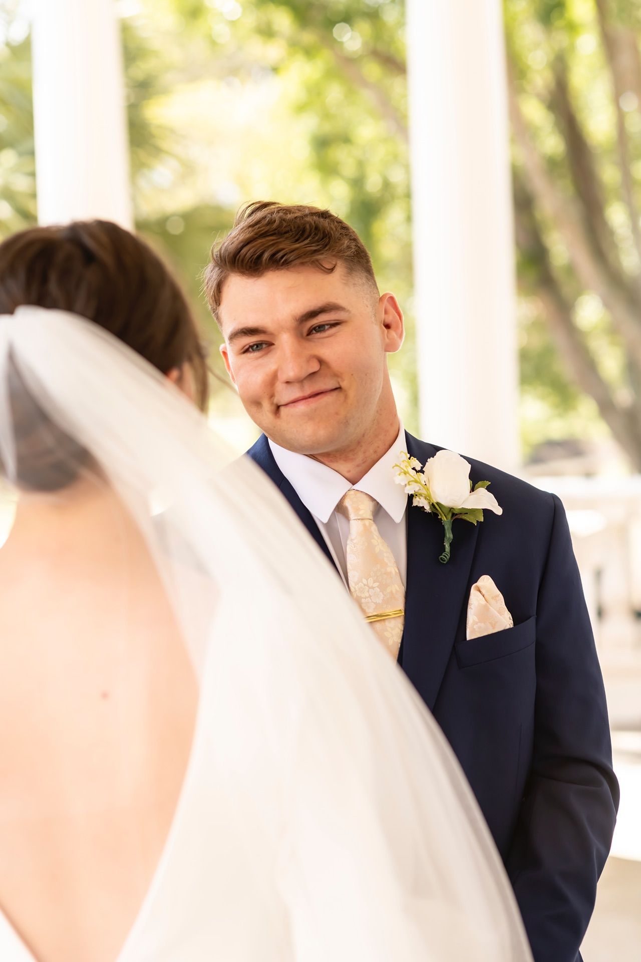 A groom in a navy suit with yellow tie smiles lovingly while looking at his bride during their wedding ceremony.