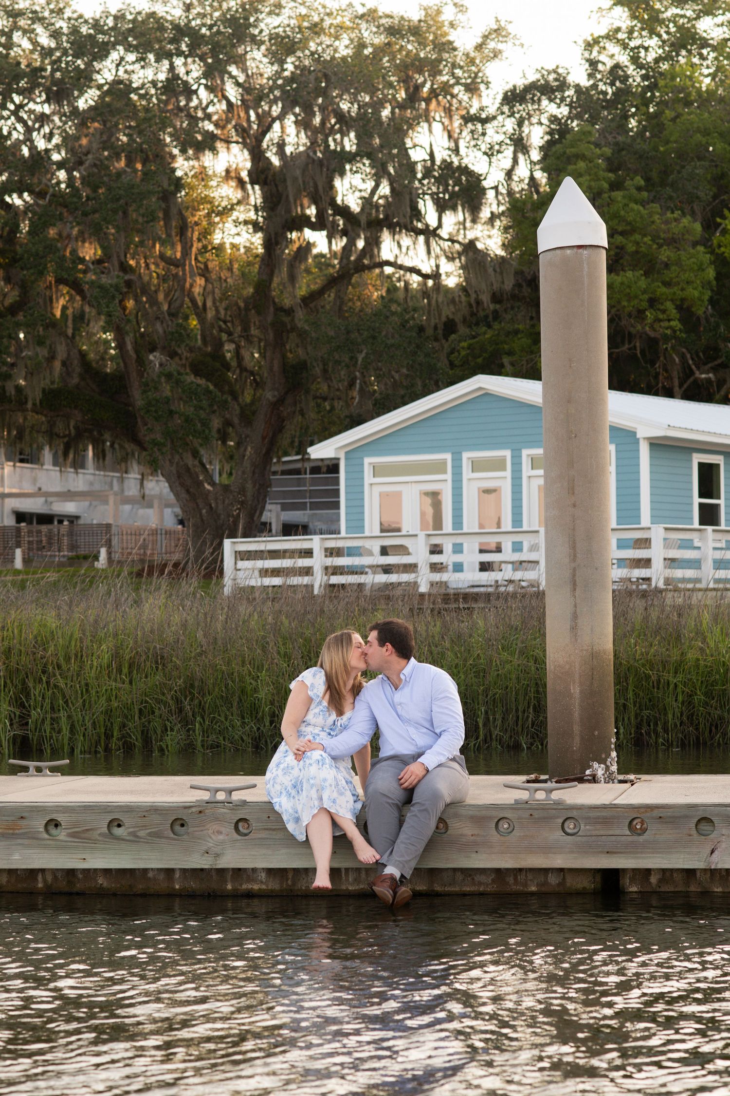 A couple sits together on a waterfront dock at sunset with a blue house and trees in the background.