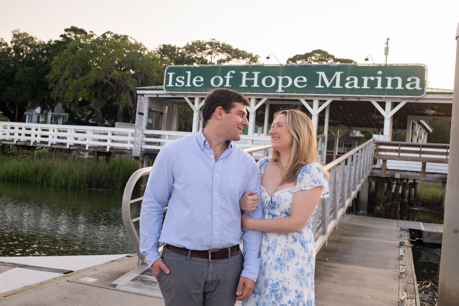 A couple in coordinating blue outfits shares a romantic moment on a wooden dock by the water during sunset.