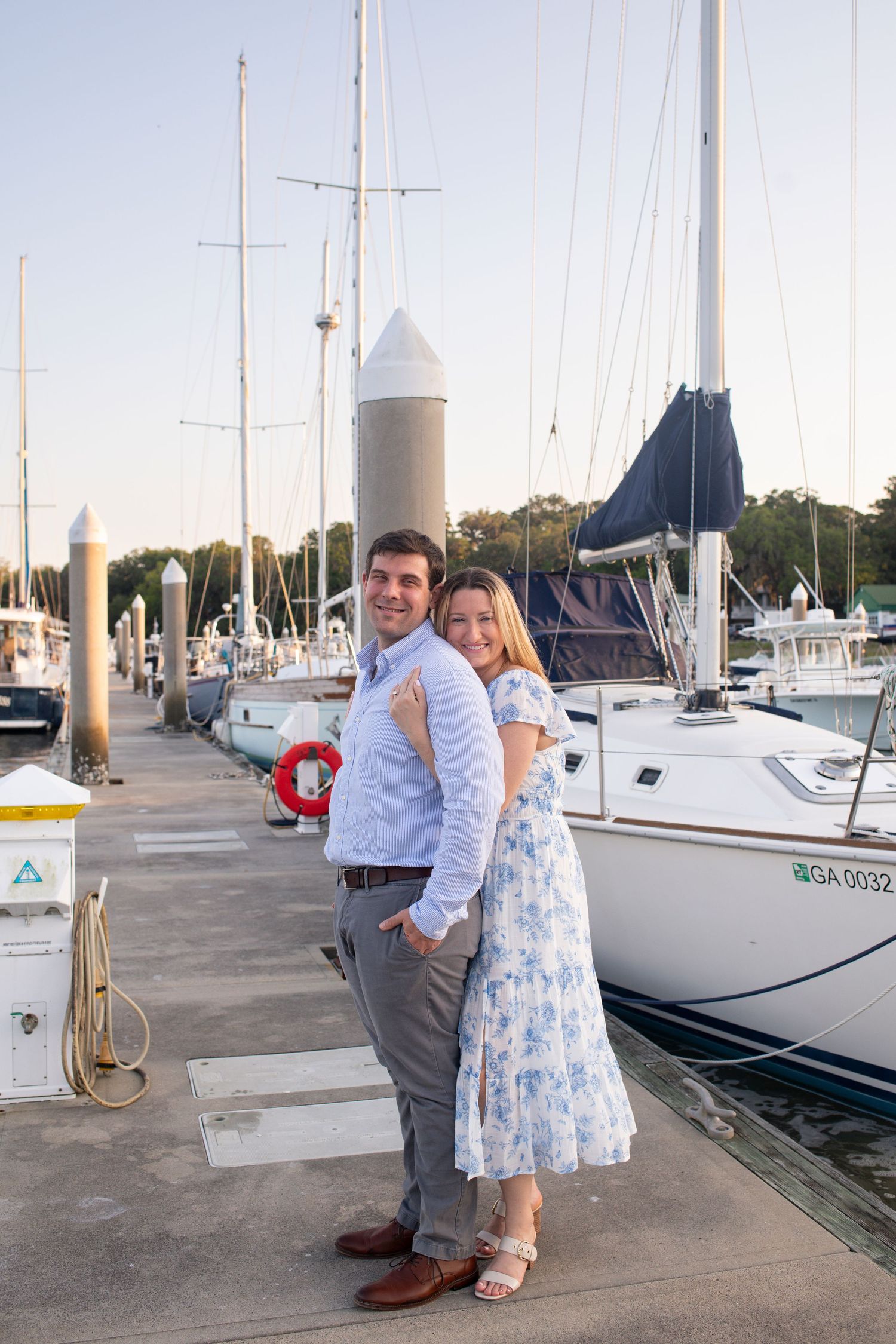 A couple poses together on a wooden dock at a marina with sailboats in the background during sunset.