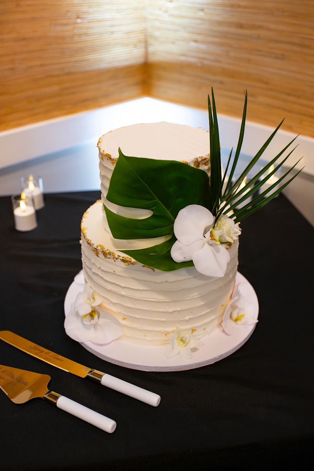 A white frosted cake decorated with tropical leaves and orchids on a black serving plate.
