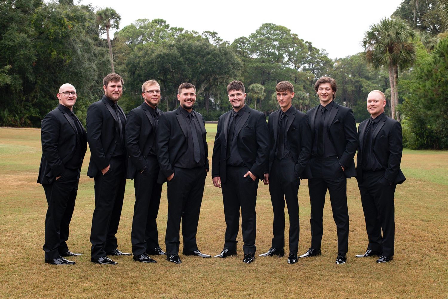 A group wearing black formal attire poses together outdoors on a grassy field.