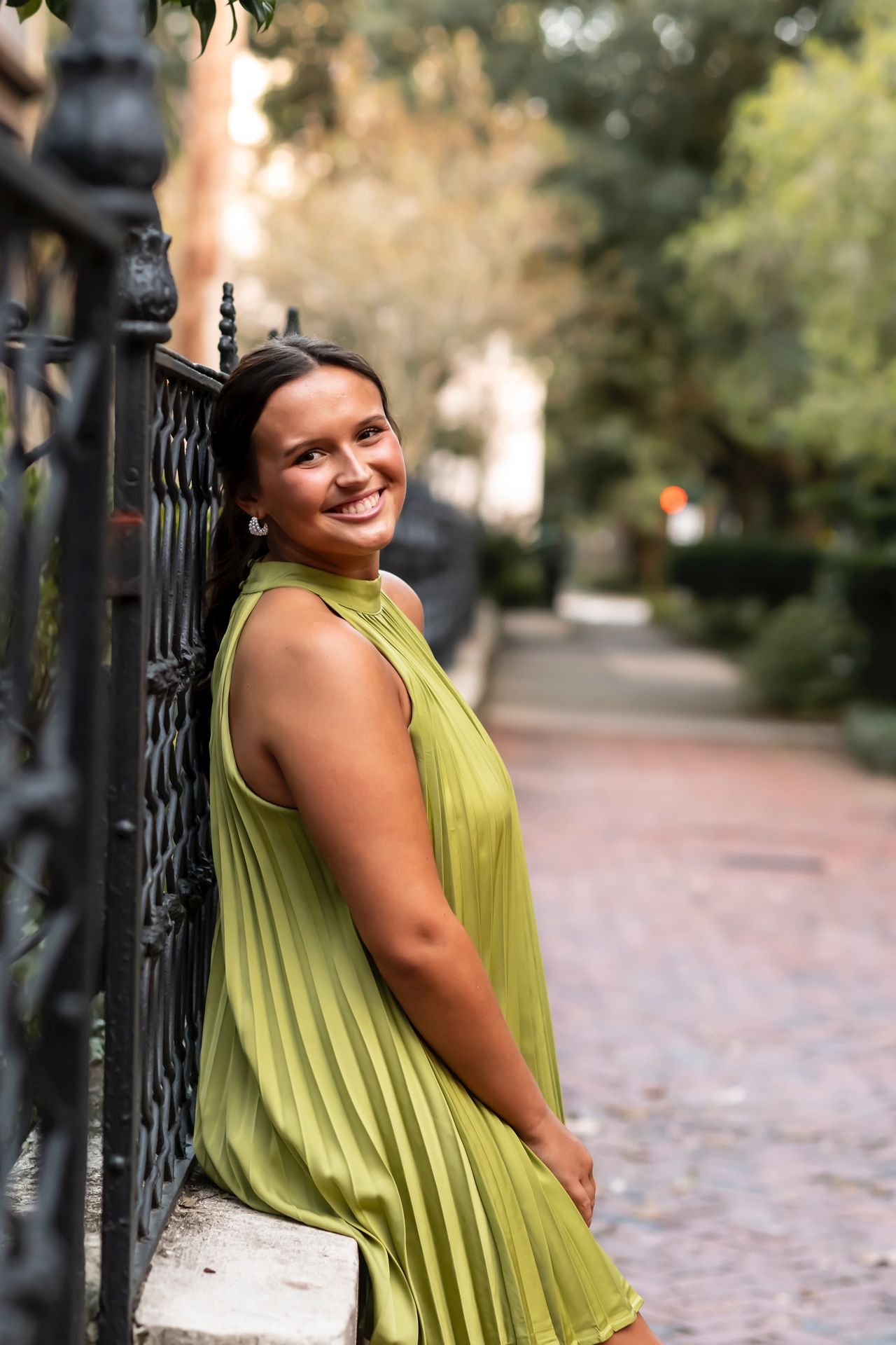 A woman in a lime green dress poses playfully against a wrought iron fence on a tree-lined urban pathway.