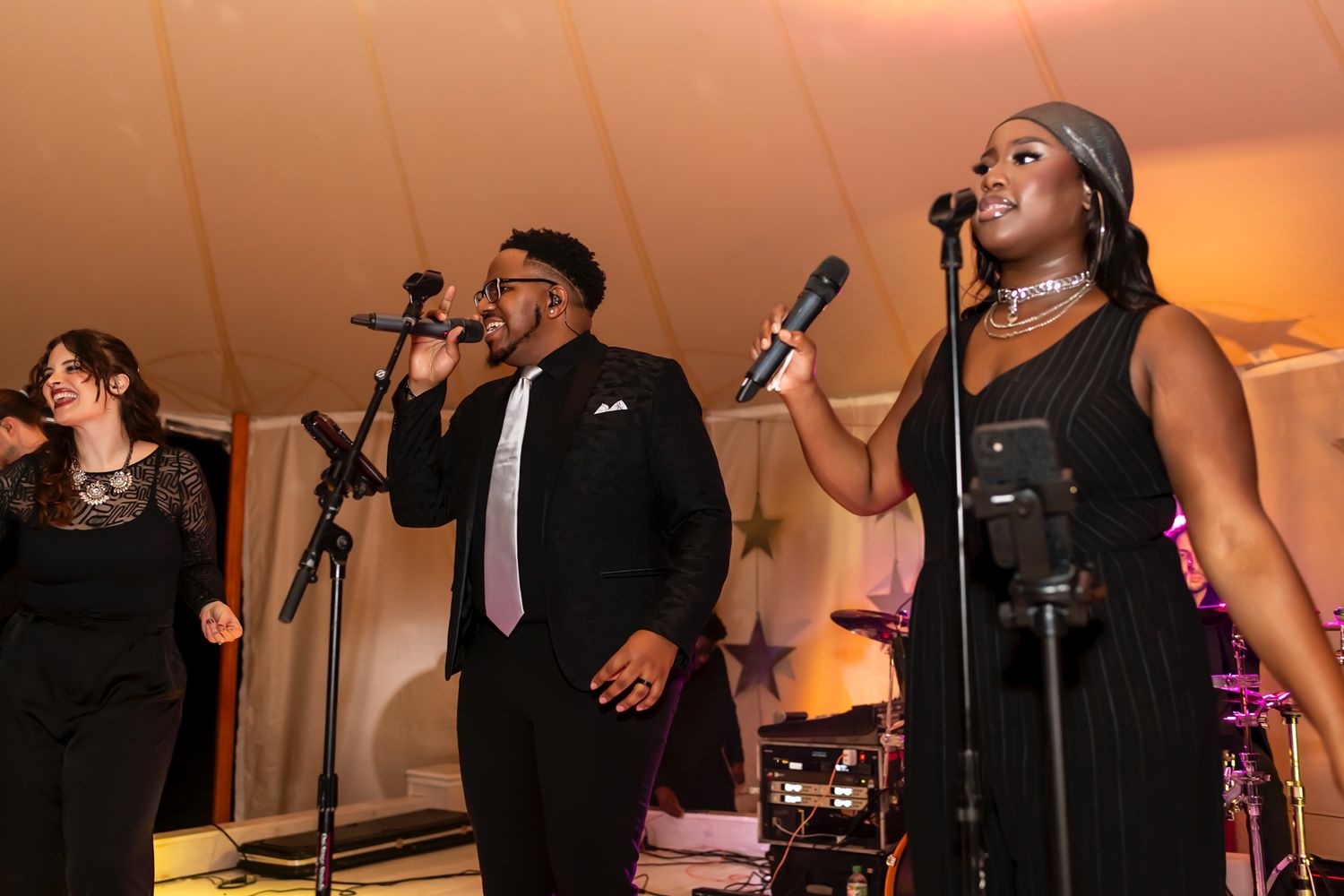 Performers sing into microphones on stage at an event with warm orange lighting and a tent-like venue setting.