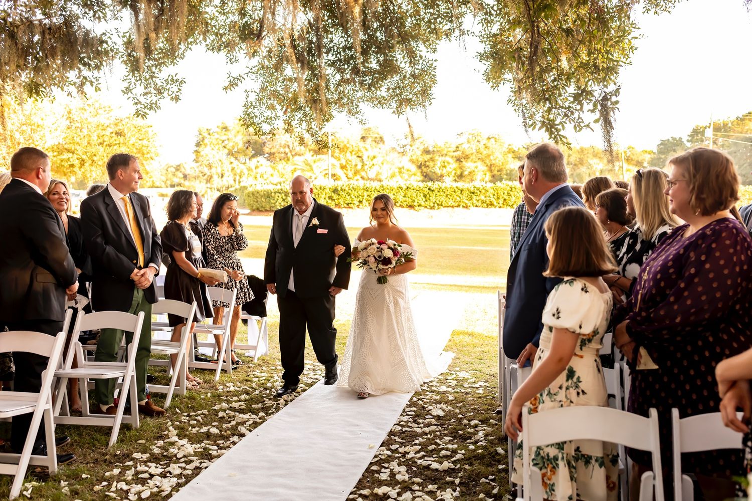 Wedding guests gather under shade trees for an outdoor ceremony during golden hour.