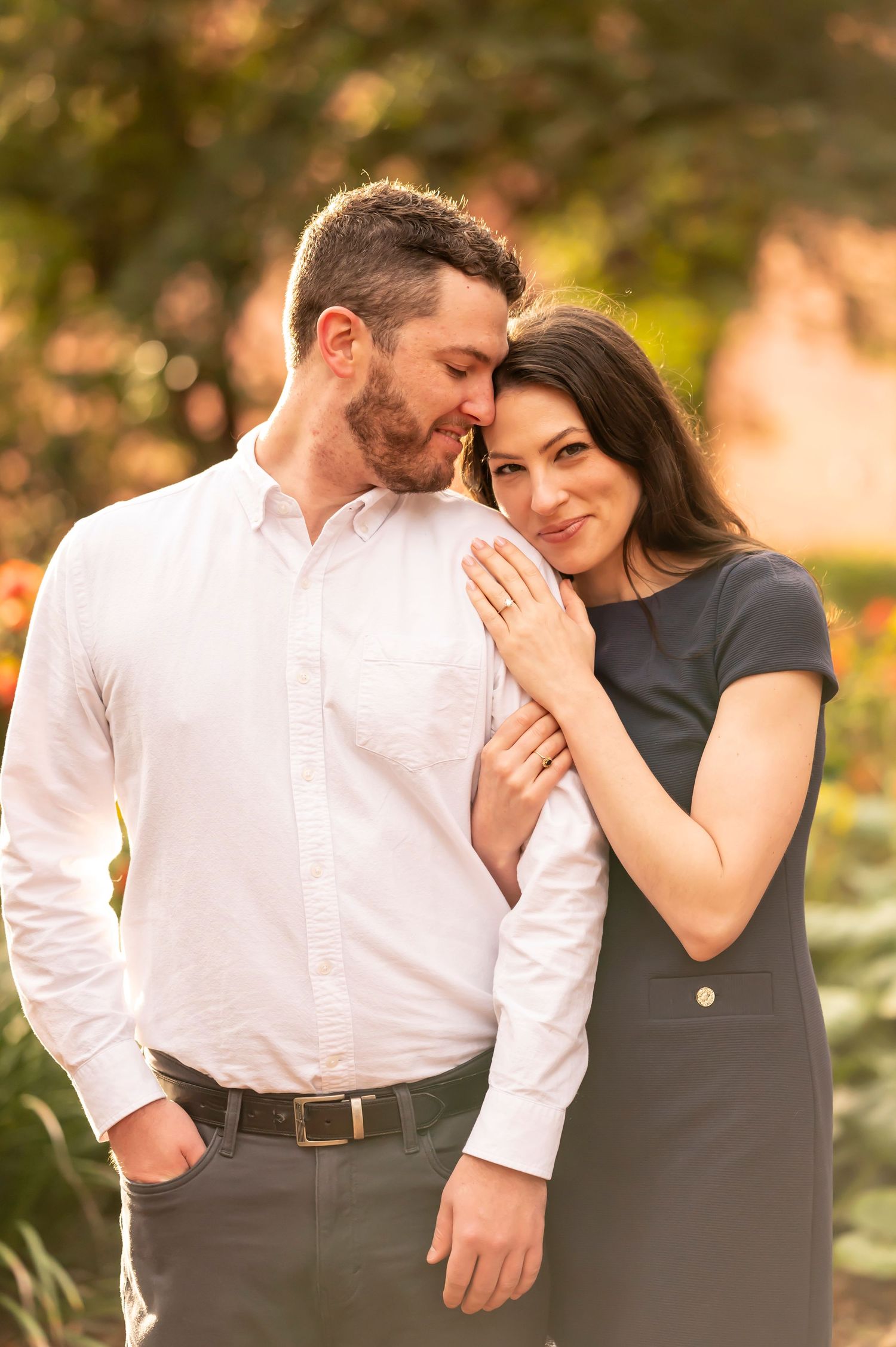 A romantic couple embracing outdoors in warm sunset light, wearing formal attire with a soft natural backdrop.