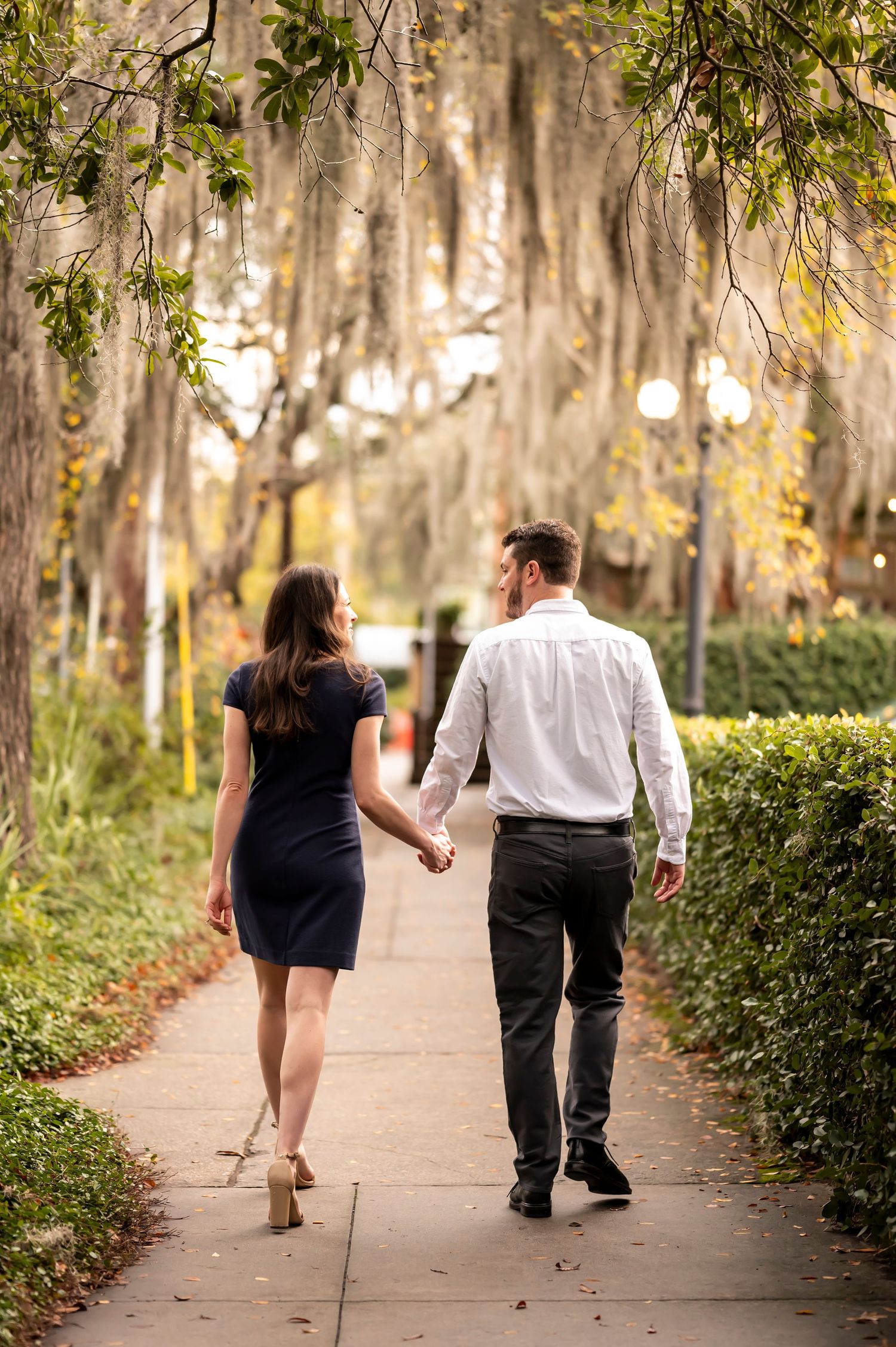 A couple walking together on a tree-lined path with Spanish moss hanging from branches above.