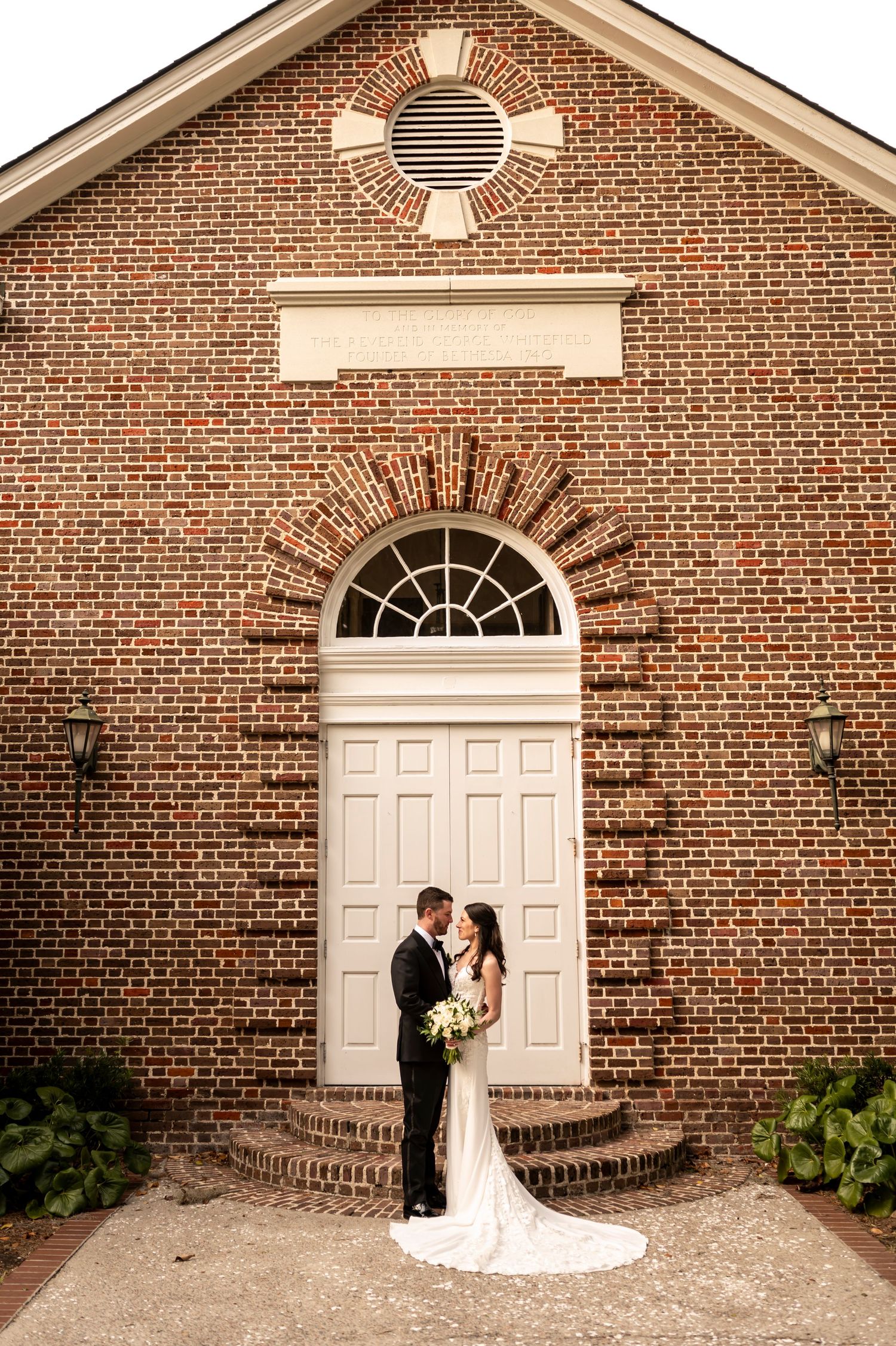Elegant wedding couple stands before grand brick church entrance with white door and circular window detail.